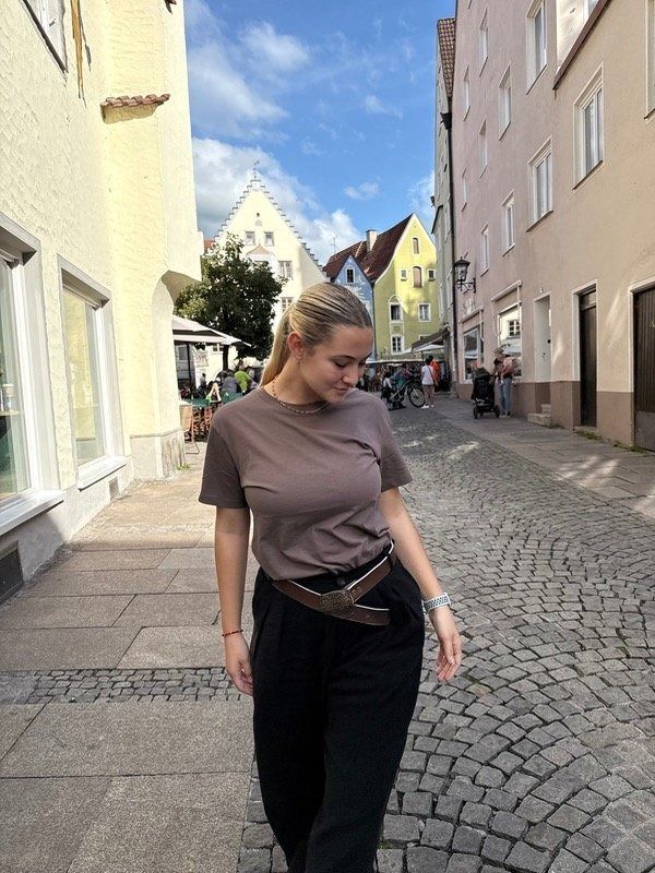 Mujer con blusa marrón y pantalones negros, manos en los bolsillos, con un cinturón marrón en una calle empedrada.