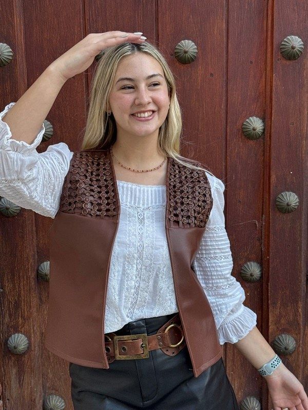 Una mujer con chaleco marrón y blusa blanca sonríe, con la mano en la cabeza, delante de una puerta de madera.