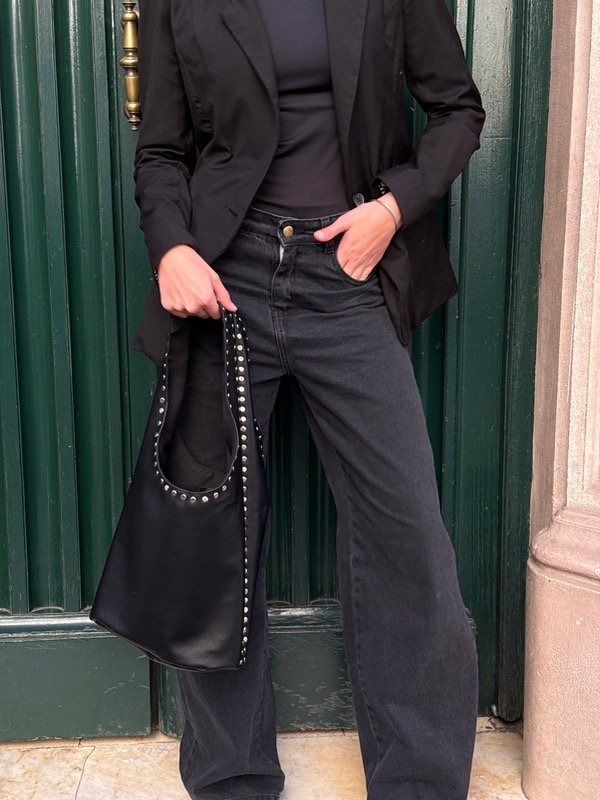 Persona vestida de negro que sostiene un bolso tote con tachuelas, de pie frente a una puerta verde.