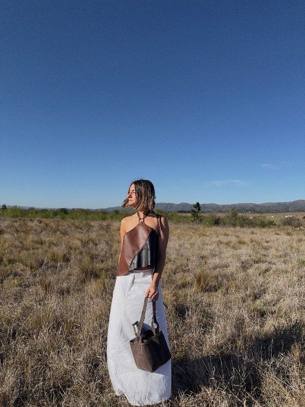 Mujer con atuendo de cuero bicolor posa cerca de una puerta de madera y una ventana arqueada con vistas a los árboles.