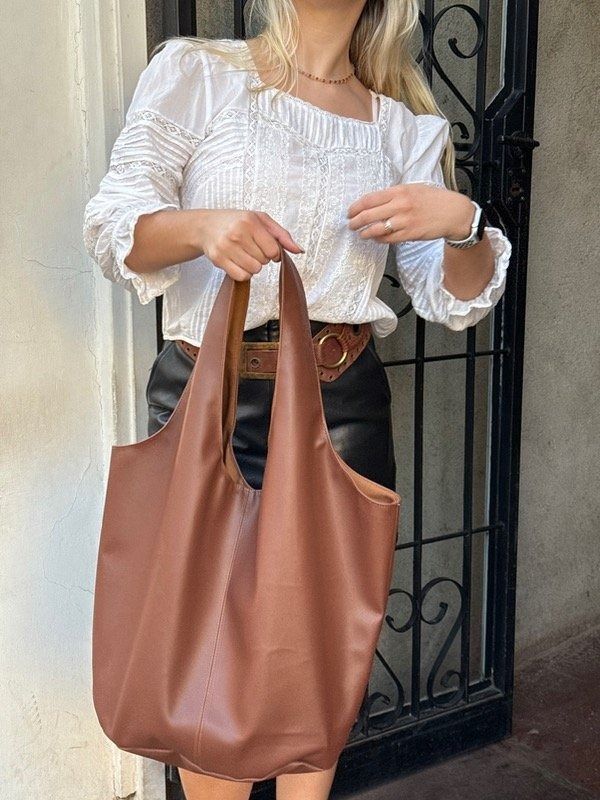 Una mujer con blusa blanca y bolso marrón levanta el brazo, sonriendo, en un pasillo de ladrillo.