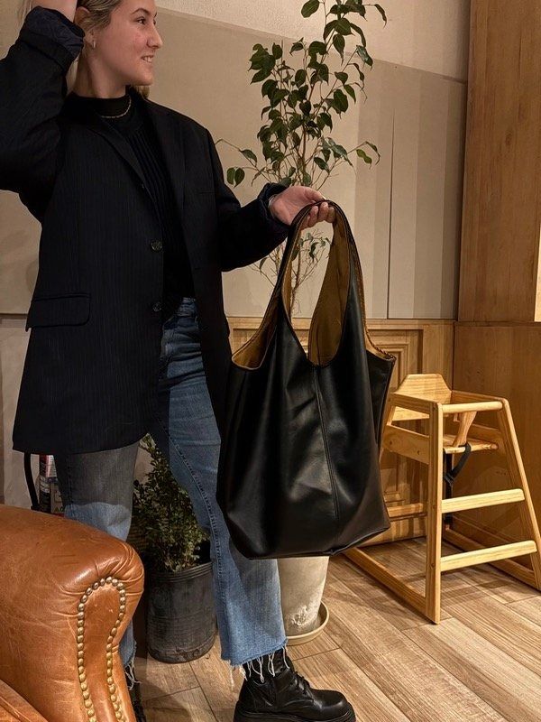 Mujer que sostiene un bolso tote negro grande, vestida con blazer, suéter y jeans en el interior.