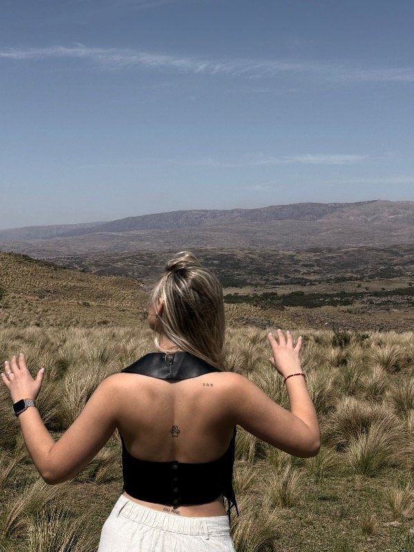 Mujer protegiéndose los ojos, de pie en un campo con una cordillera al fondo, cielo azul.
