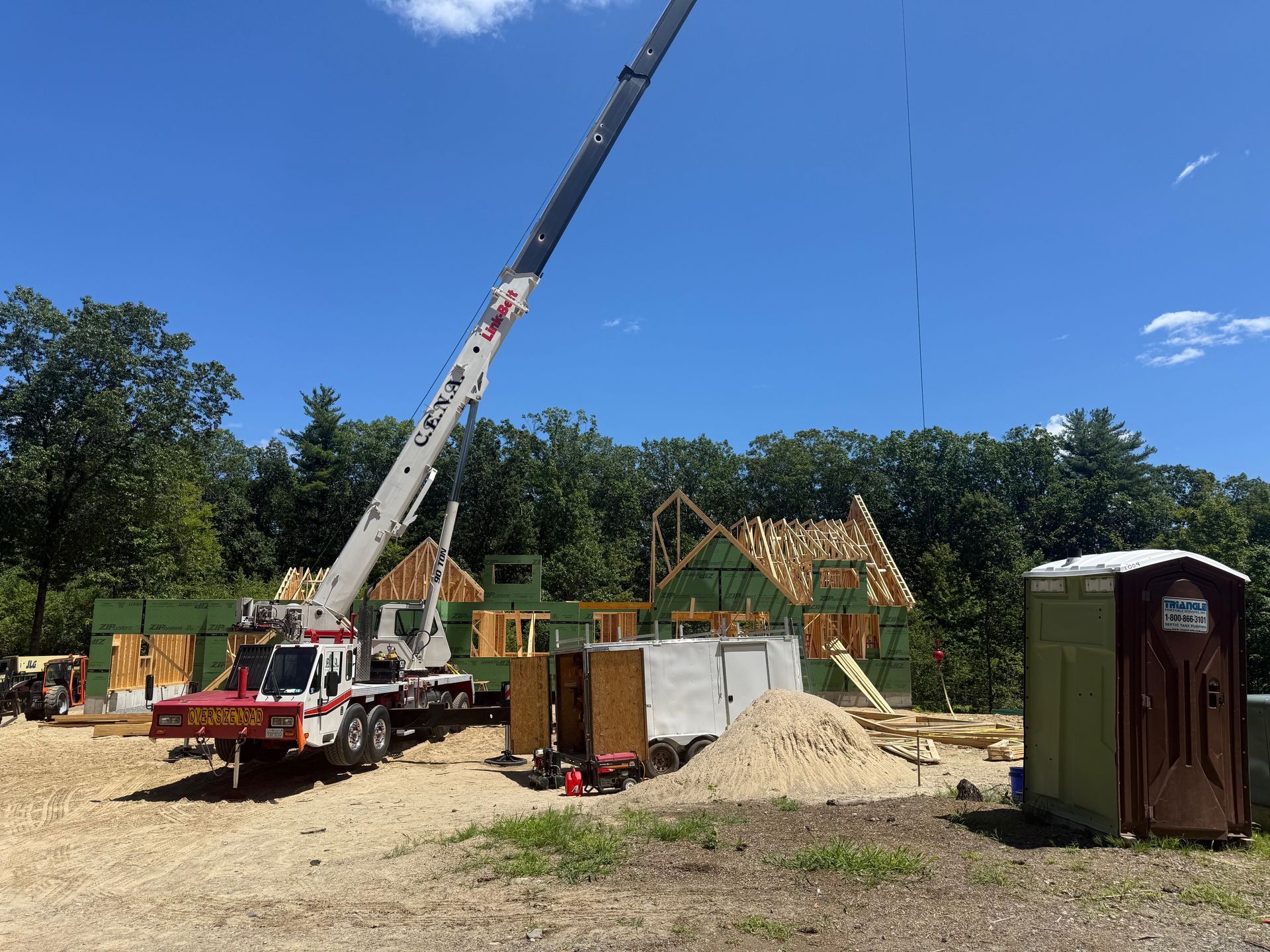 Crane lifting lumber at a construction site with partially built wooden frame house, blue sky, and portable toilet.