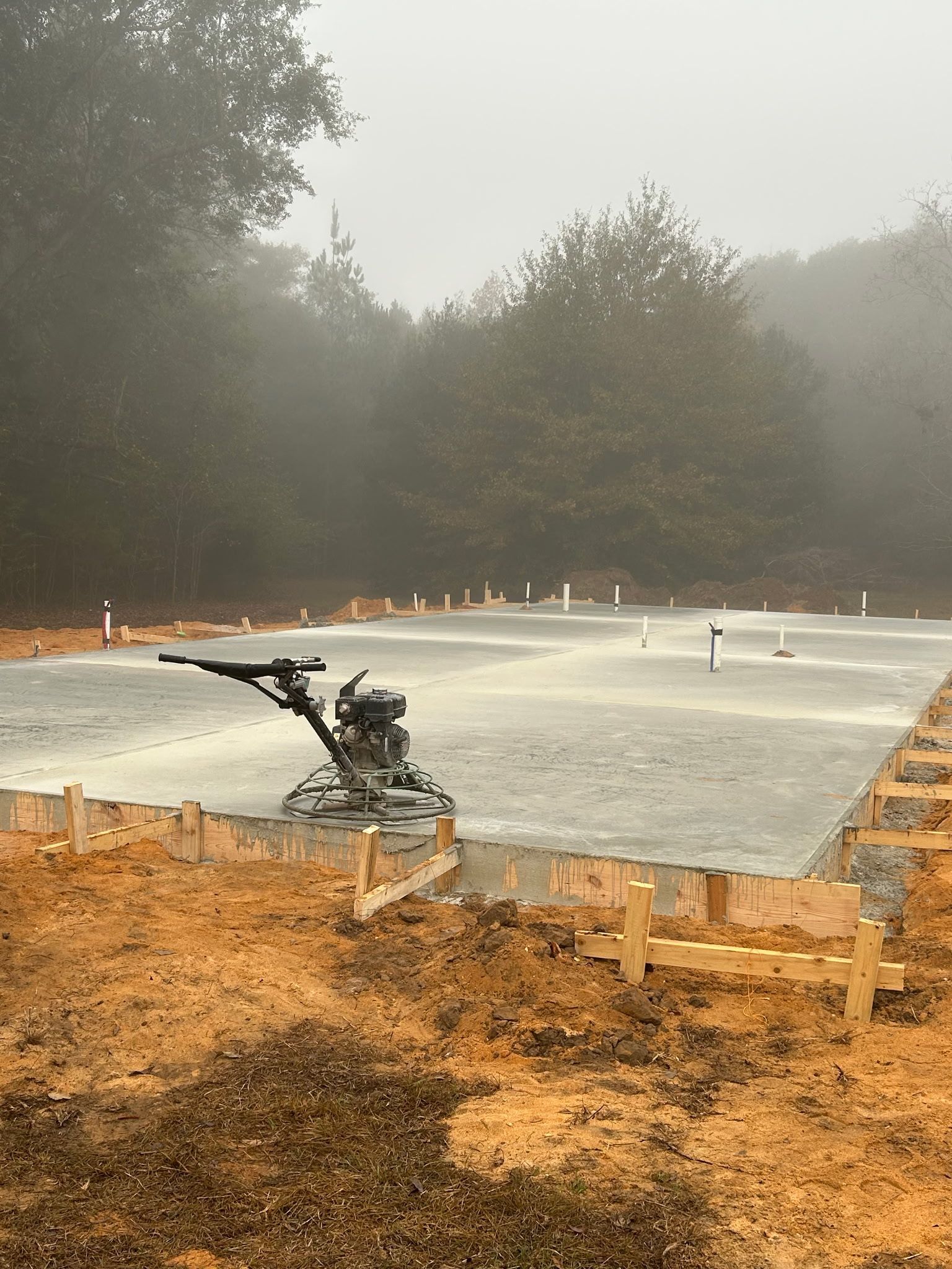 Newly poured concrete slab with a power trowel, surrounded by wooden framing and bare earth, in a misty forest.