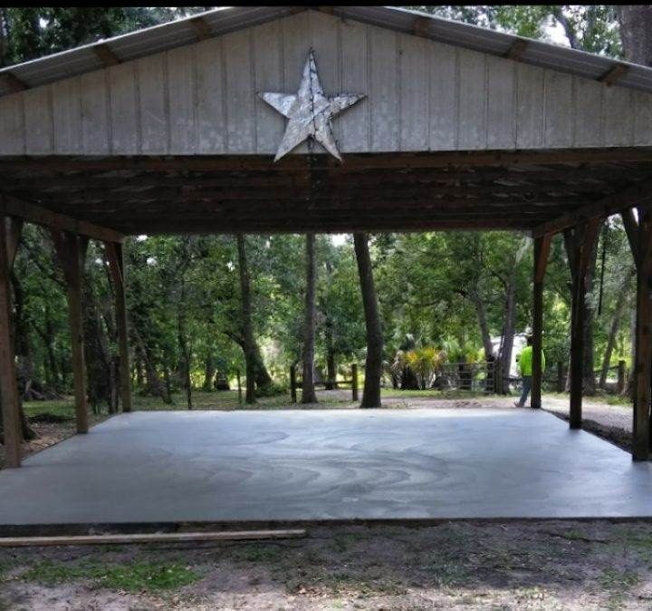 Open-air concrete-floored pavilion in a wooded area with a metal roof and a large star decoration.