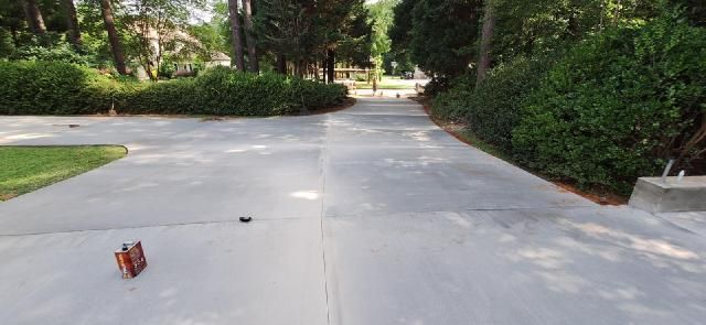A concrete driveway leads toward trees, with greenery on either side. A small red box sits on the left.