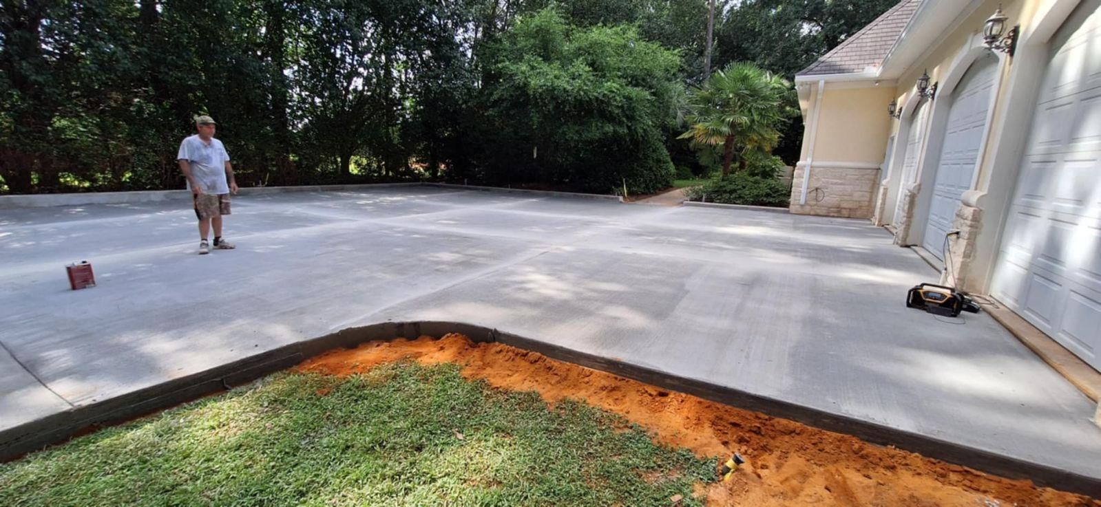 A man stands on a concrete driveway, near grass edging.  A house with garage doors is to the right.