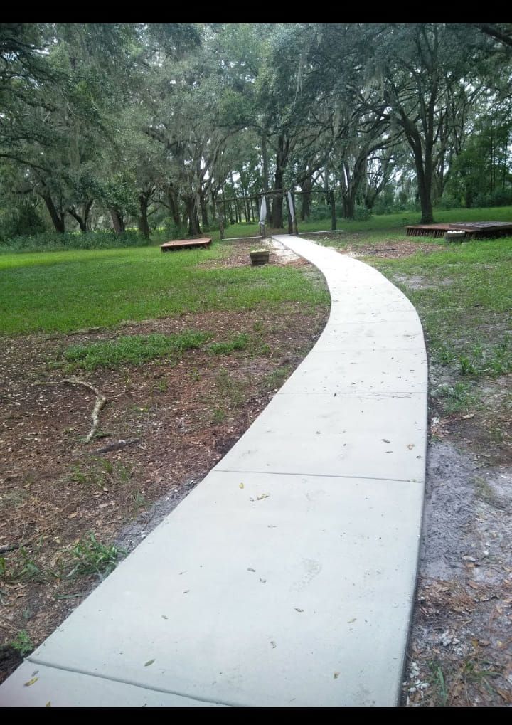 Concrete path curves through a grassy park, leading towards trees under an overcast sky.