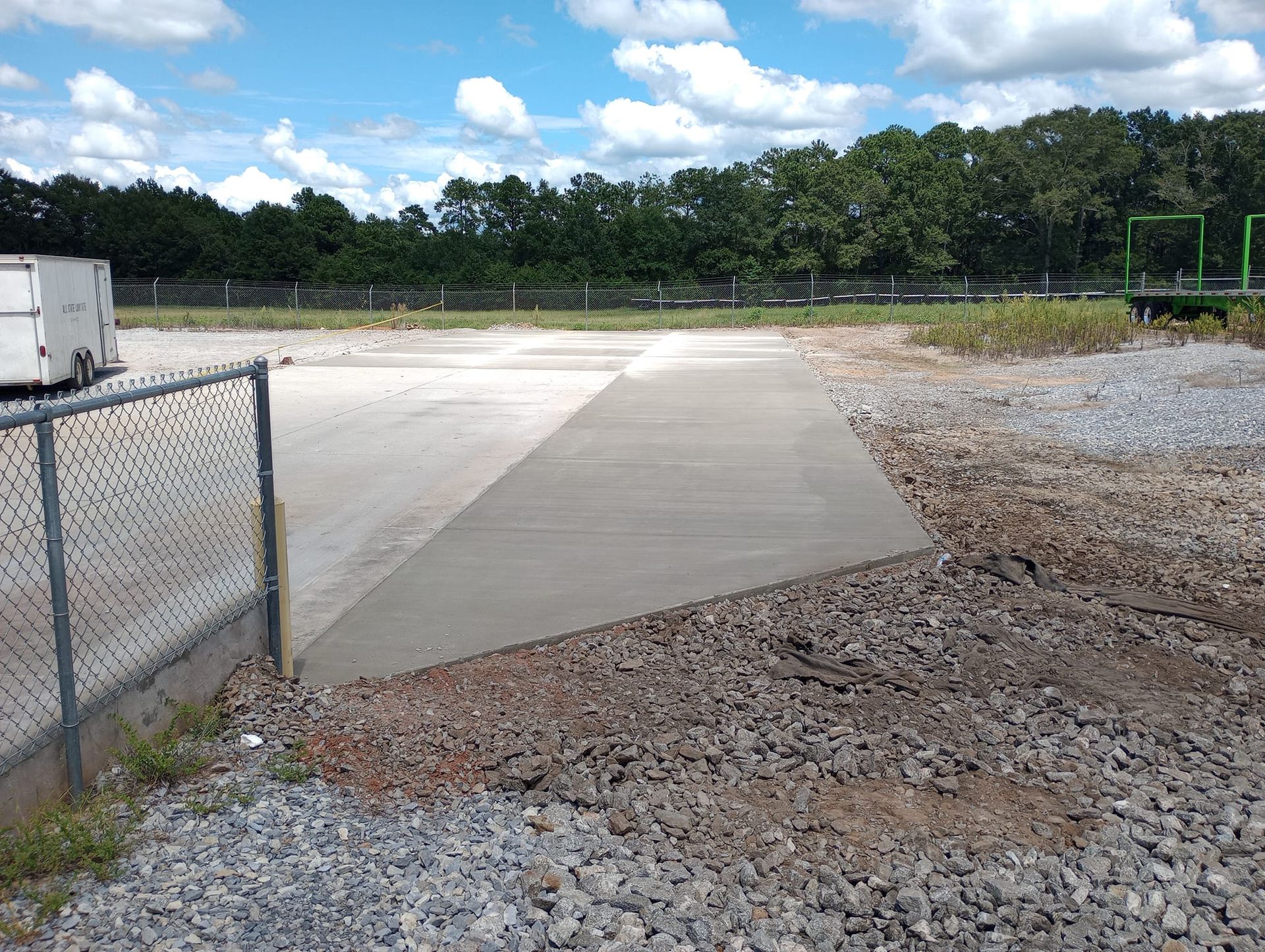 Concrete ramp leading to a parking area, surrounded by gravel and a chain-link fence. Blue sky and trees in the background.