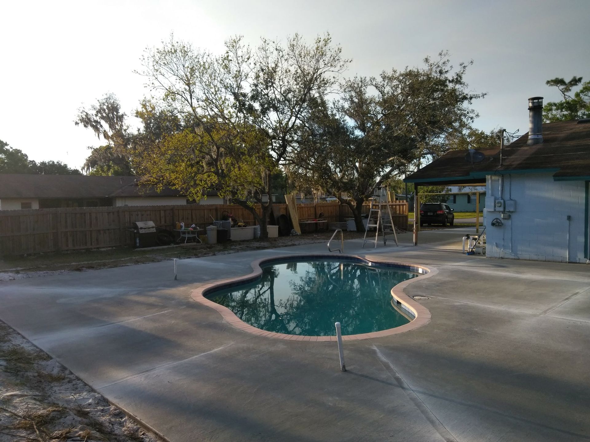Backyard with a kidney-shaped pool, concrete patio, trees, and a house.