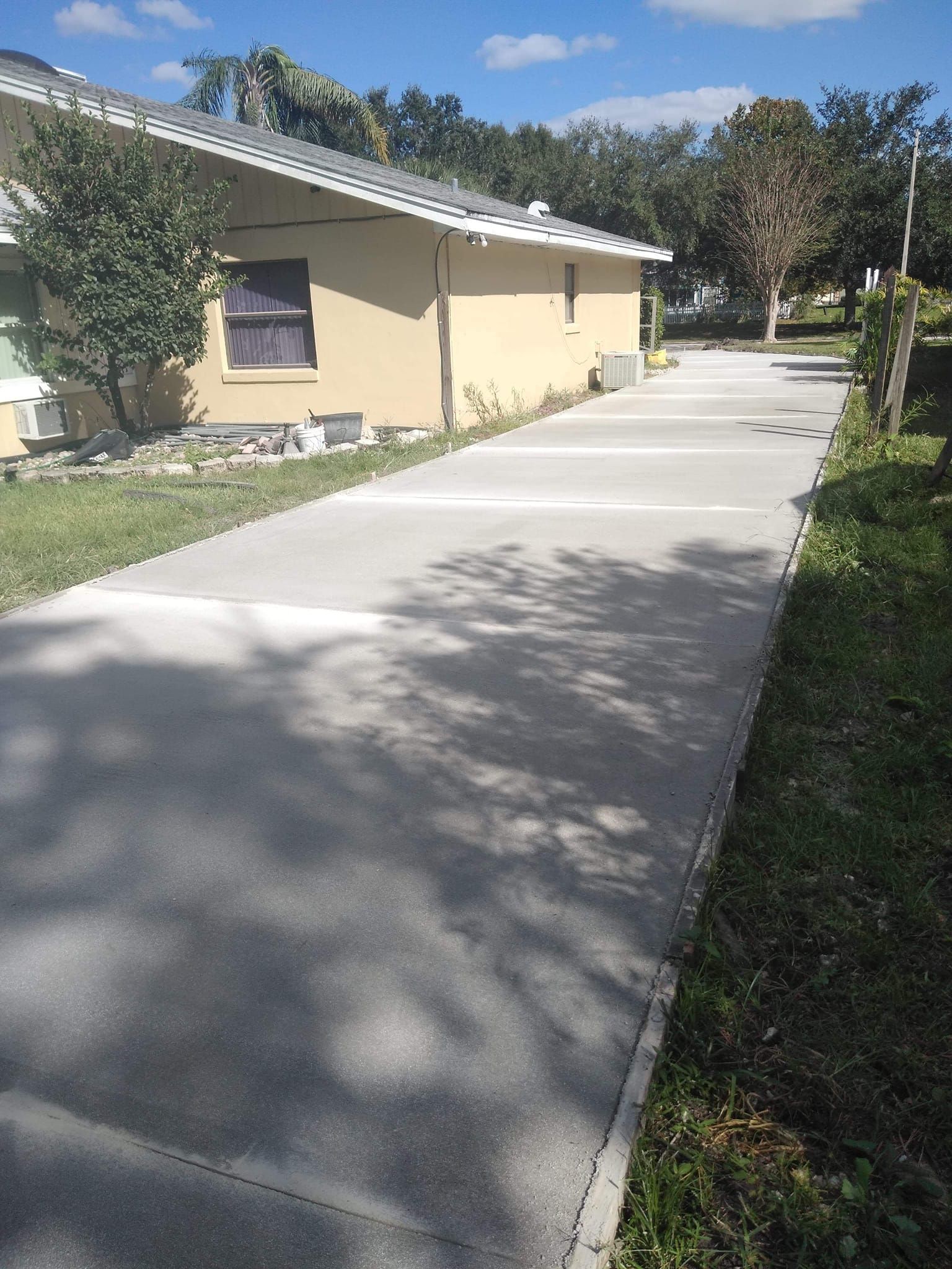 Concrete driveway alongside a tan building with a blue sky overhead. Green grass and trees are nearby.