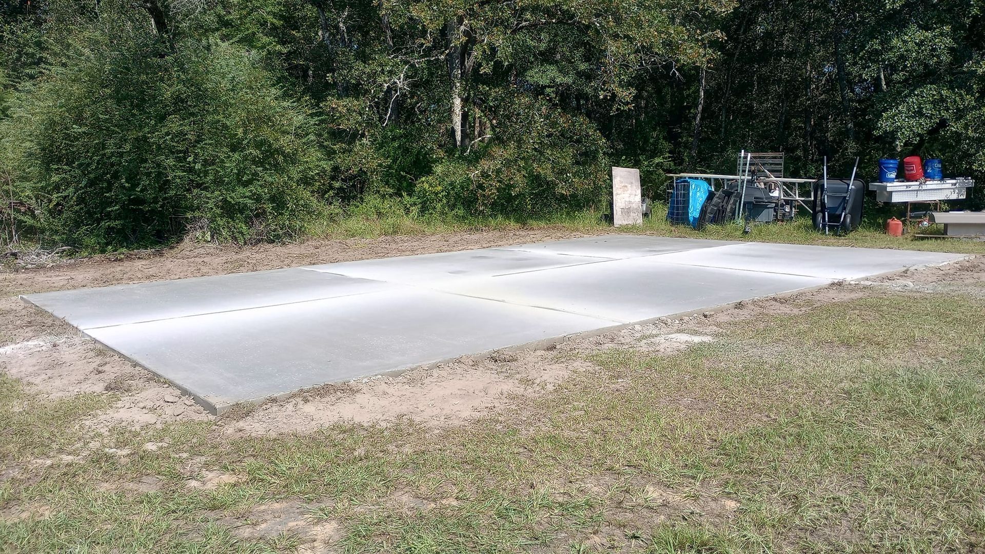 Concrete slab on grass, near trees. Blue bikes, table, supplies are visible in the background.