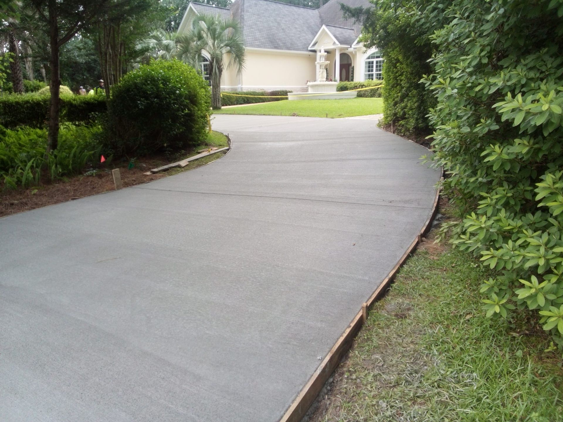 Newly paved concrete driveway leading to a beige house, bordered by green grass and bushes.