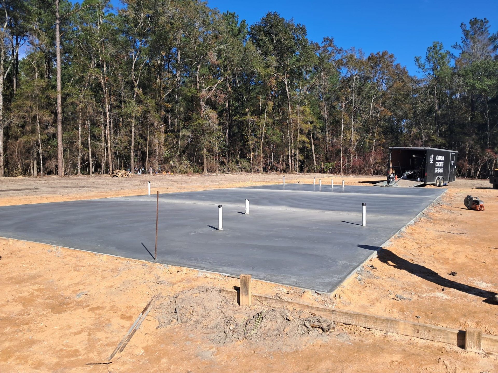 Newly poured, dark gray concrete slab with white PVC pipes, in a clearing. Background: trees and dirt.