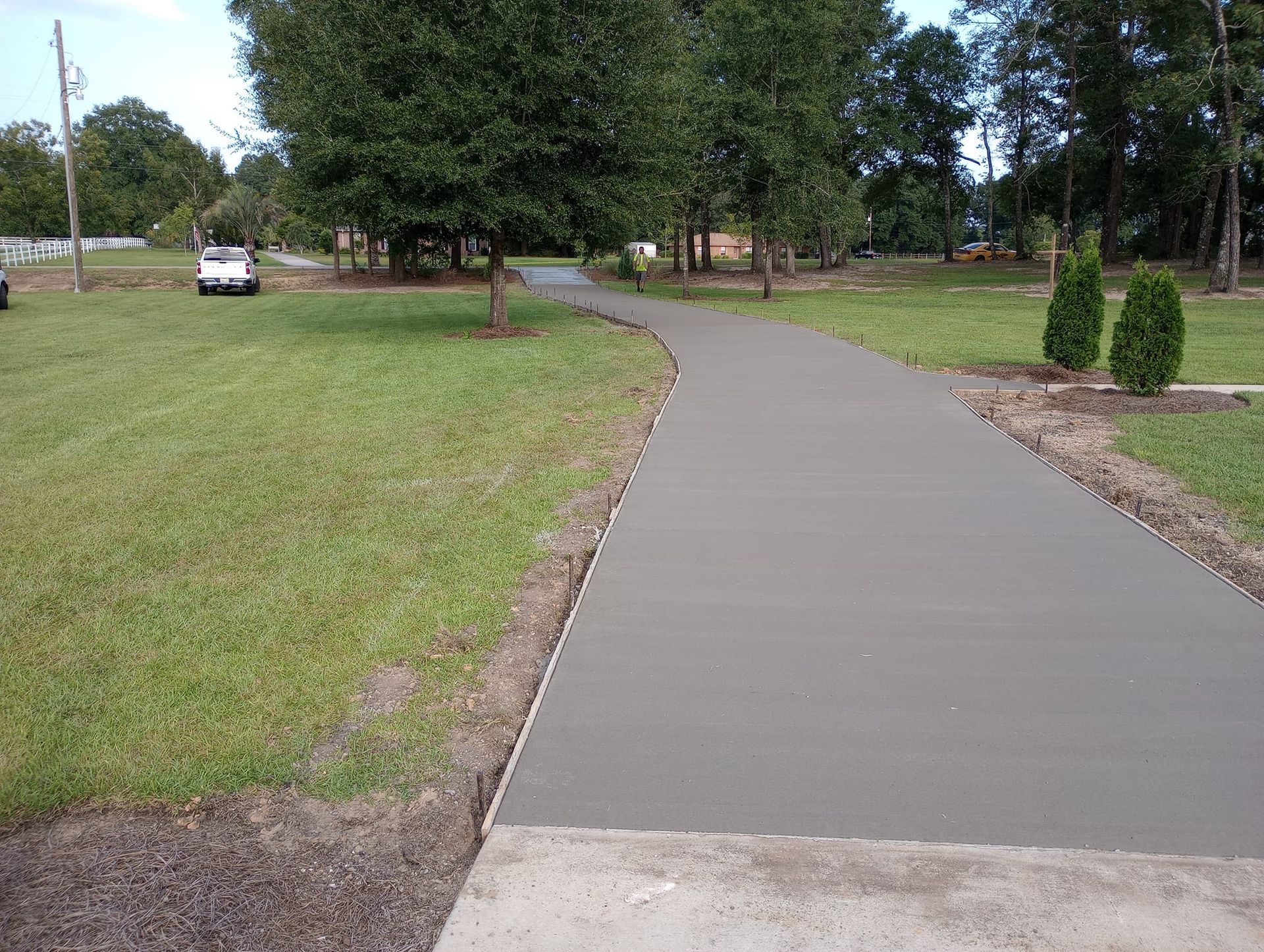 Newly poured concrete driveway curves through a grassy yard toward a distant tree line.