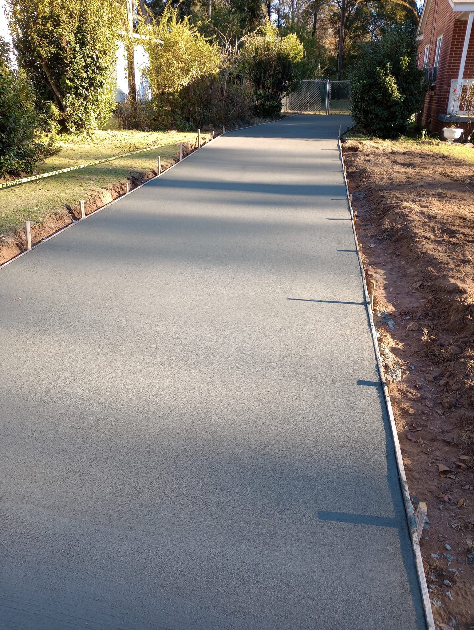 Newly poured concrete driveway, bordered by wooden forms and soil, leading towards houses in a sunny setting.