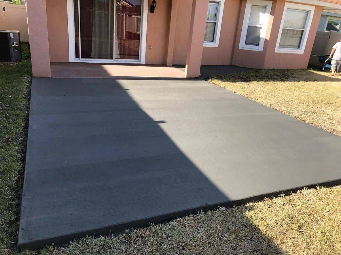 Gray concrete patio outside a light-colored house. Patio has a rectangular shape and borders grass.