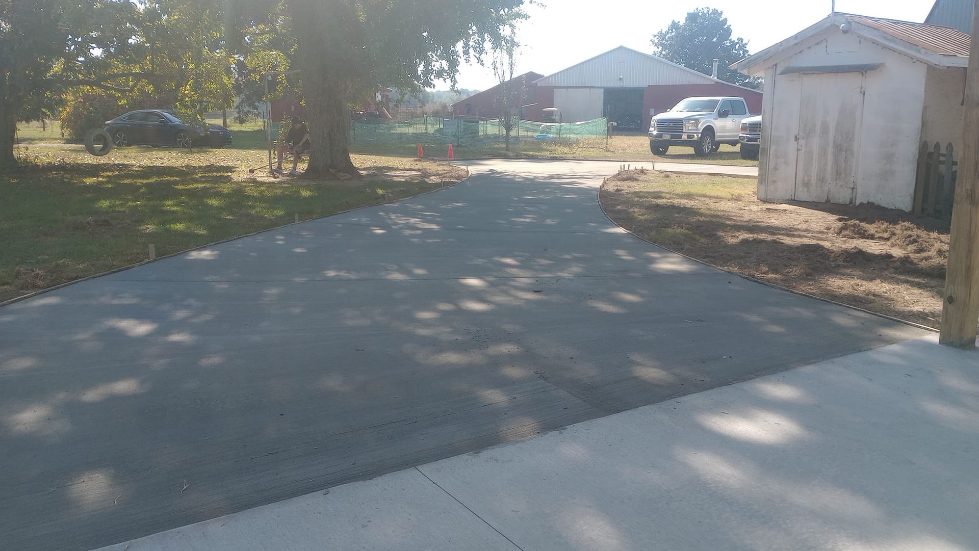 Asphalt driveway leading to a rural yard with a white truck, shed, and buildings in the background.
