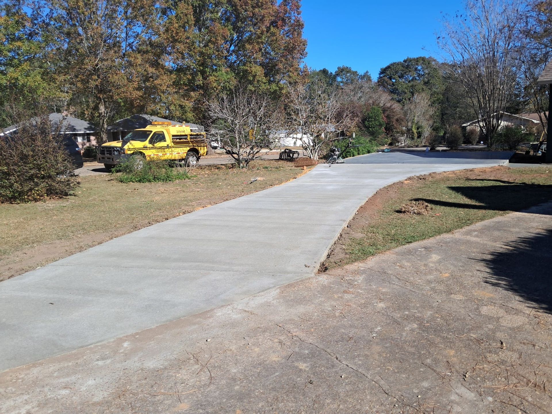 New concrete driveway curves into a residential area, with a yellow truck and trees in the background.