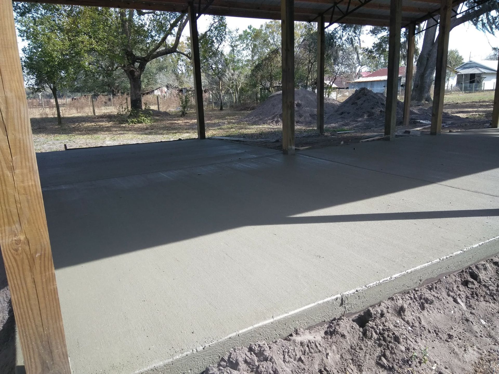 Freshly poured concrete floor under a wooden shelter, with a surrounding dirt area and trees in the background.