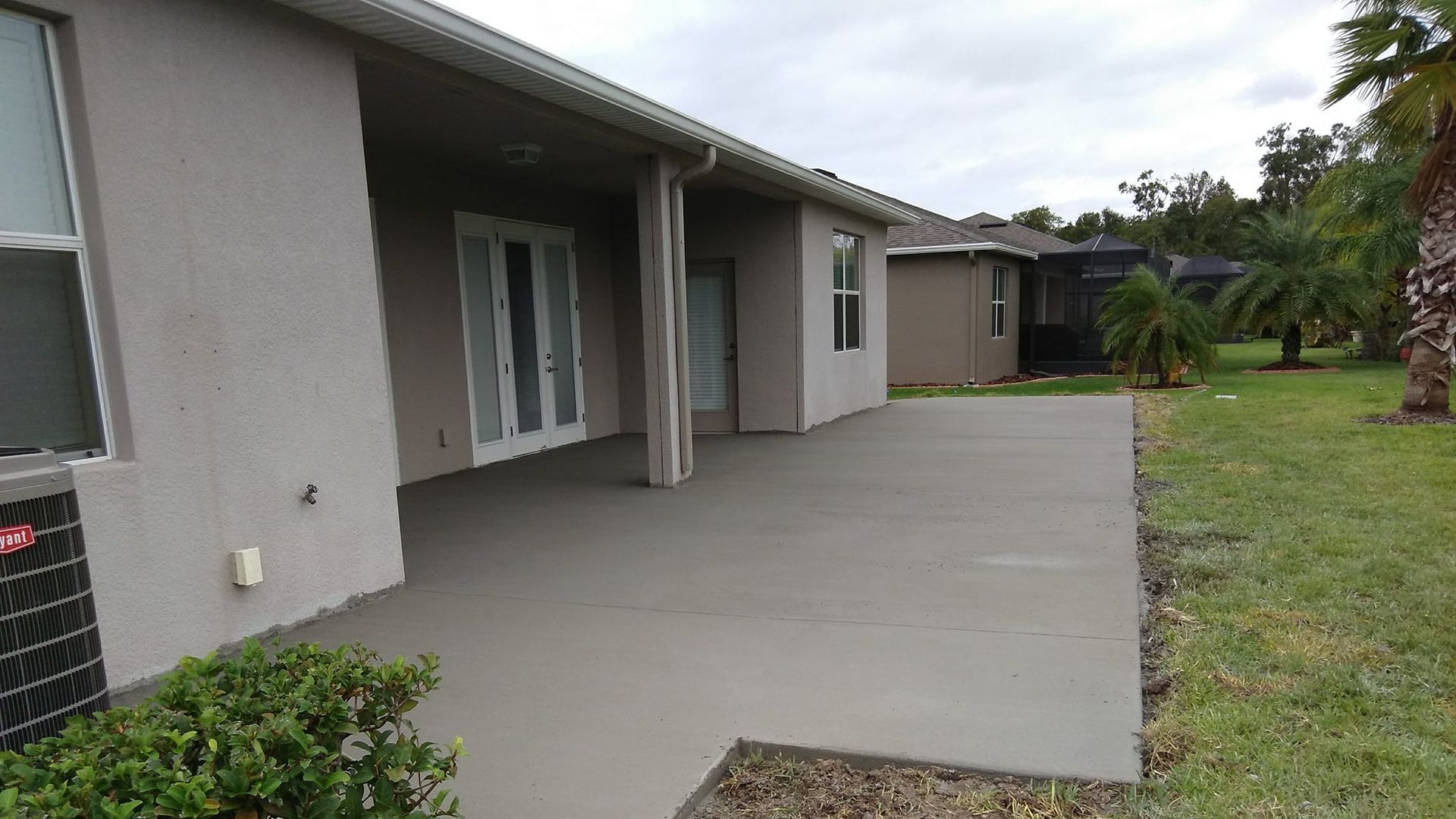 Back patio with gray concrete, adjacent to a light gray house and green lawn. Overcast sky.