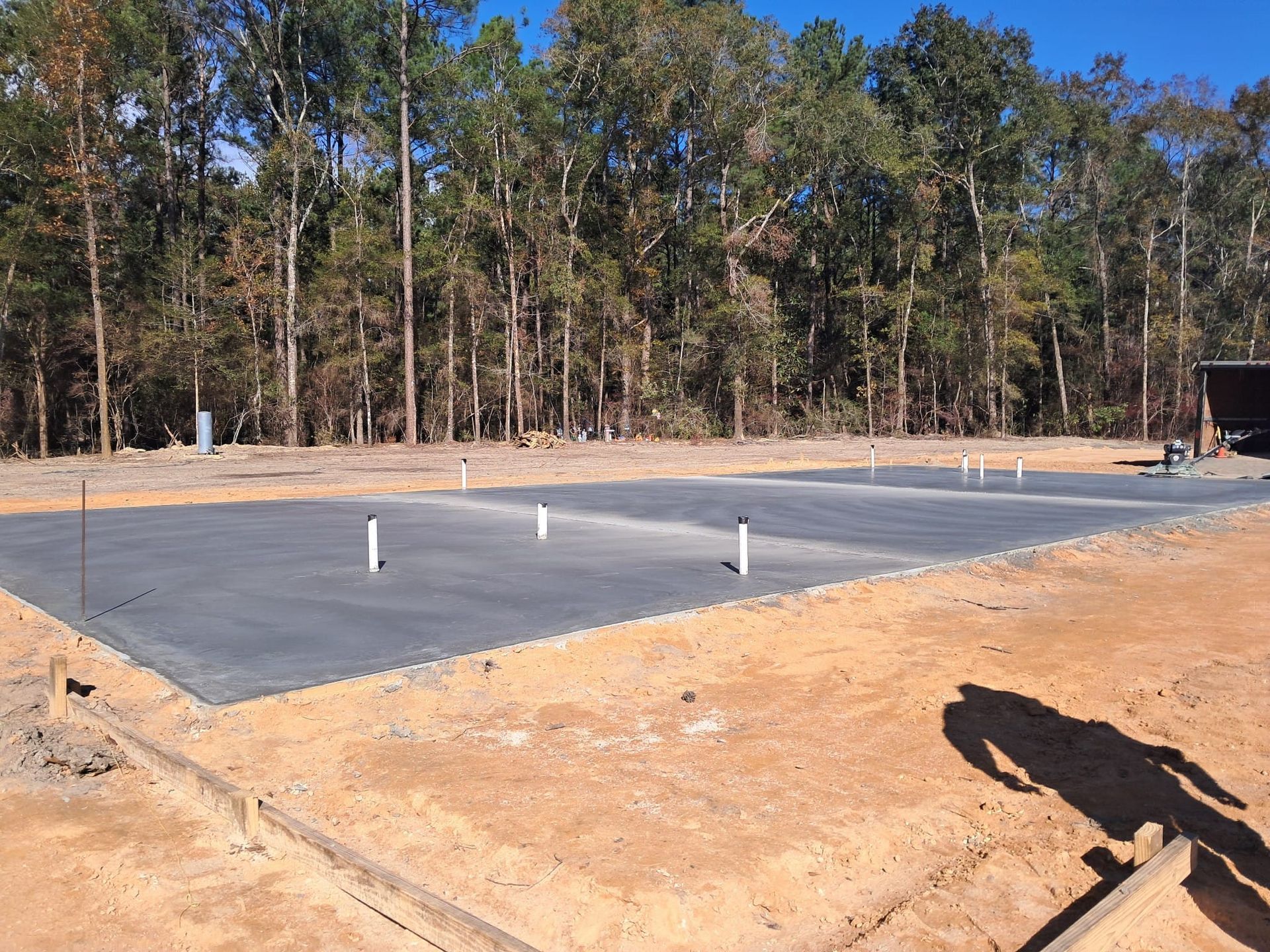 A concrete foundation with white plumbing pipes, outdoors. Trees in the background, shadow in foreground.