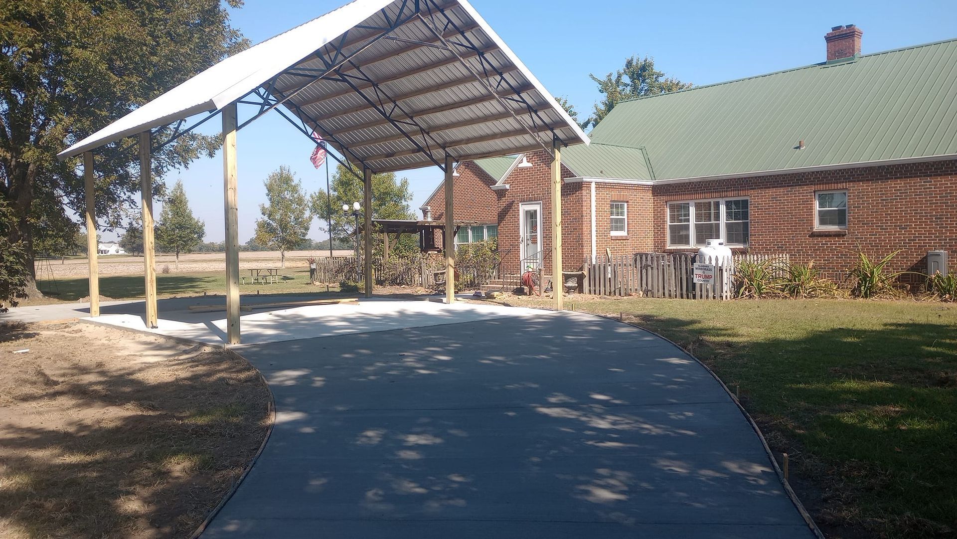 Carport over a driveway leading to a brick house with a green roof.
