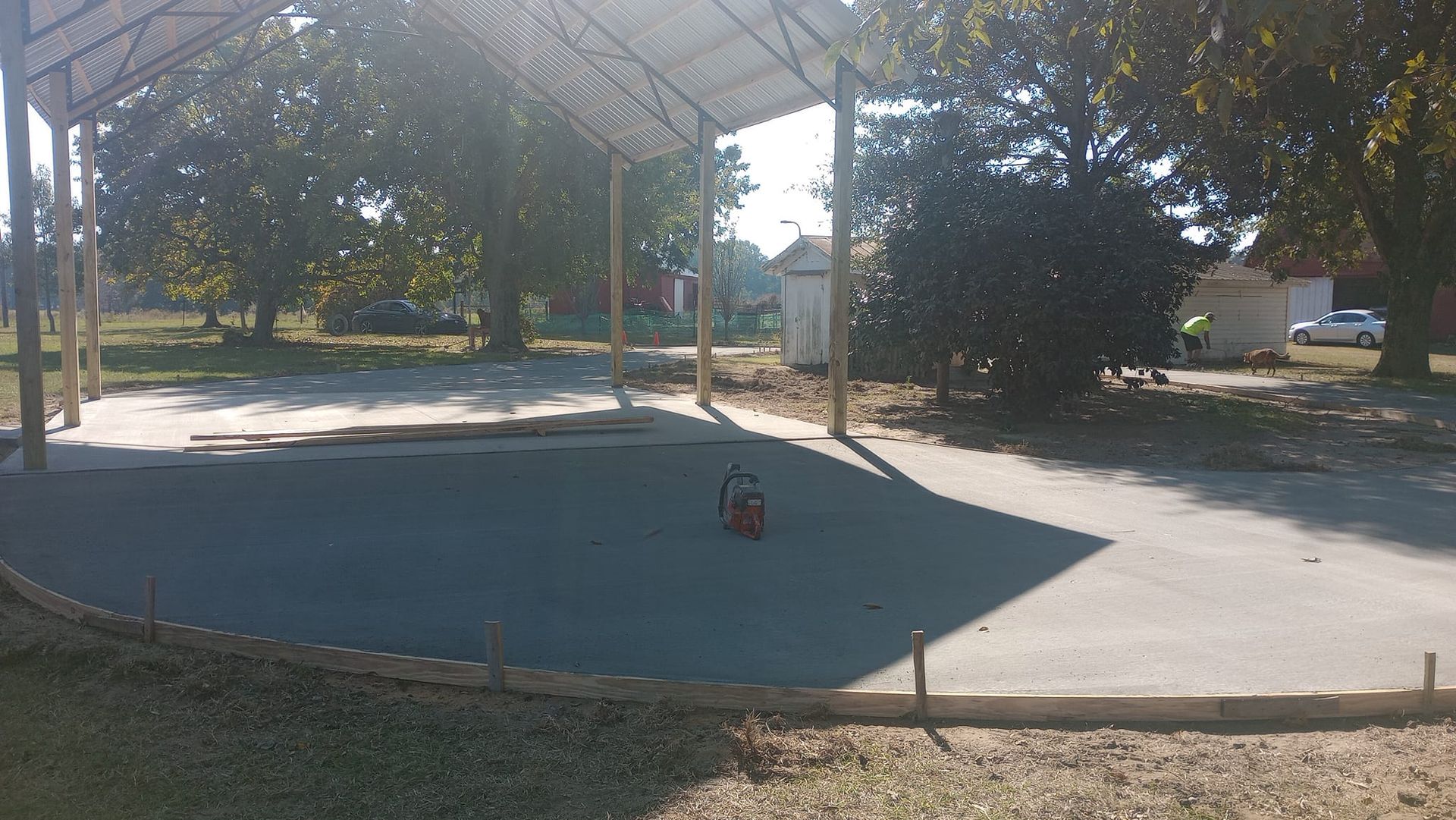 Newly poured concrete slab under a metal-framed shelter, surrounded by trees.