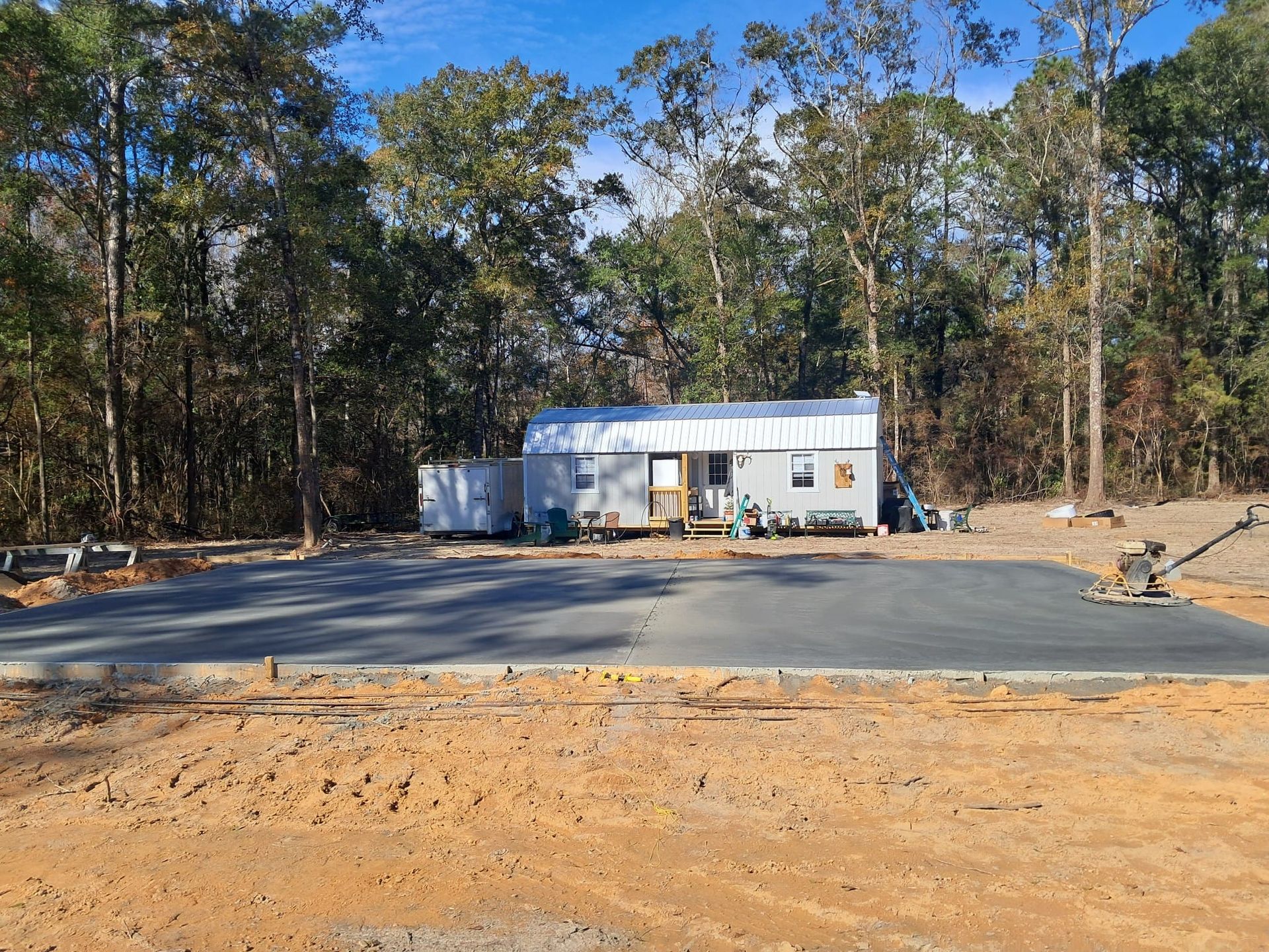 Small, rectangular building with light-colored walls and a metal roof, set on a gravel lot near a wooded area.