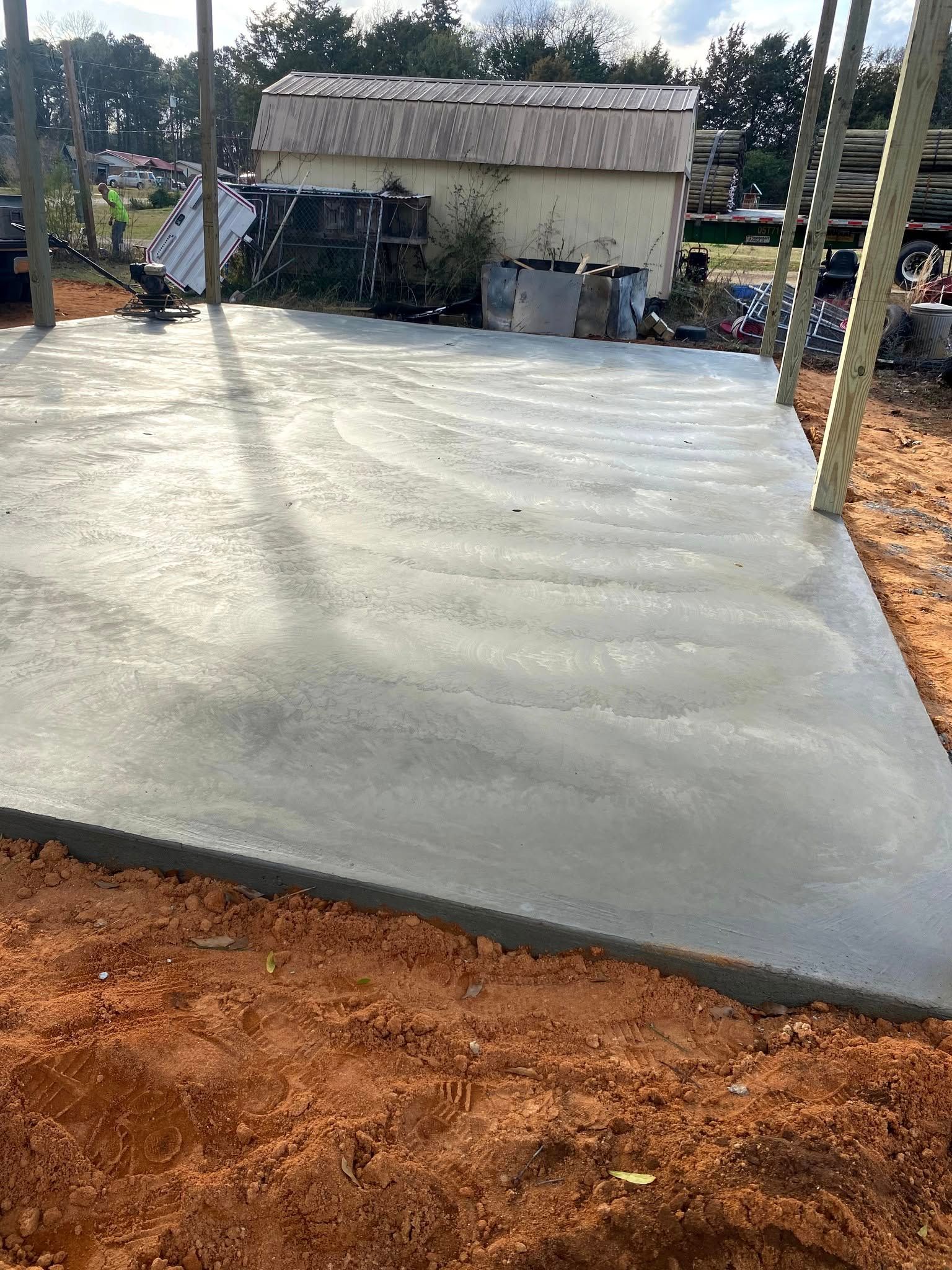 Newly poured concrete slab in front of a weathered building, surrounded by dirt and wooden posts.