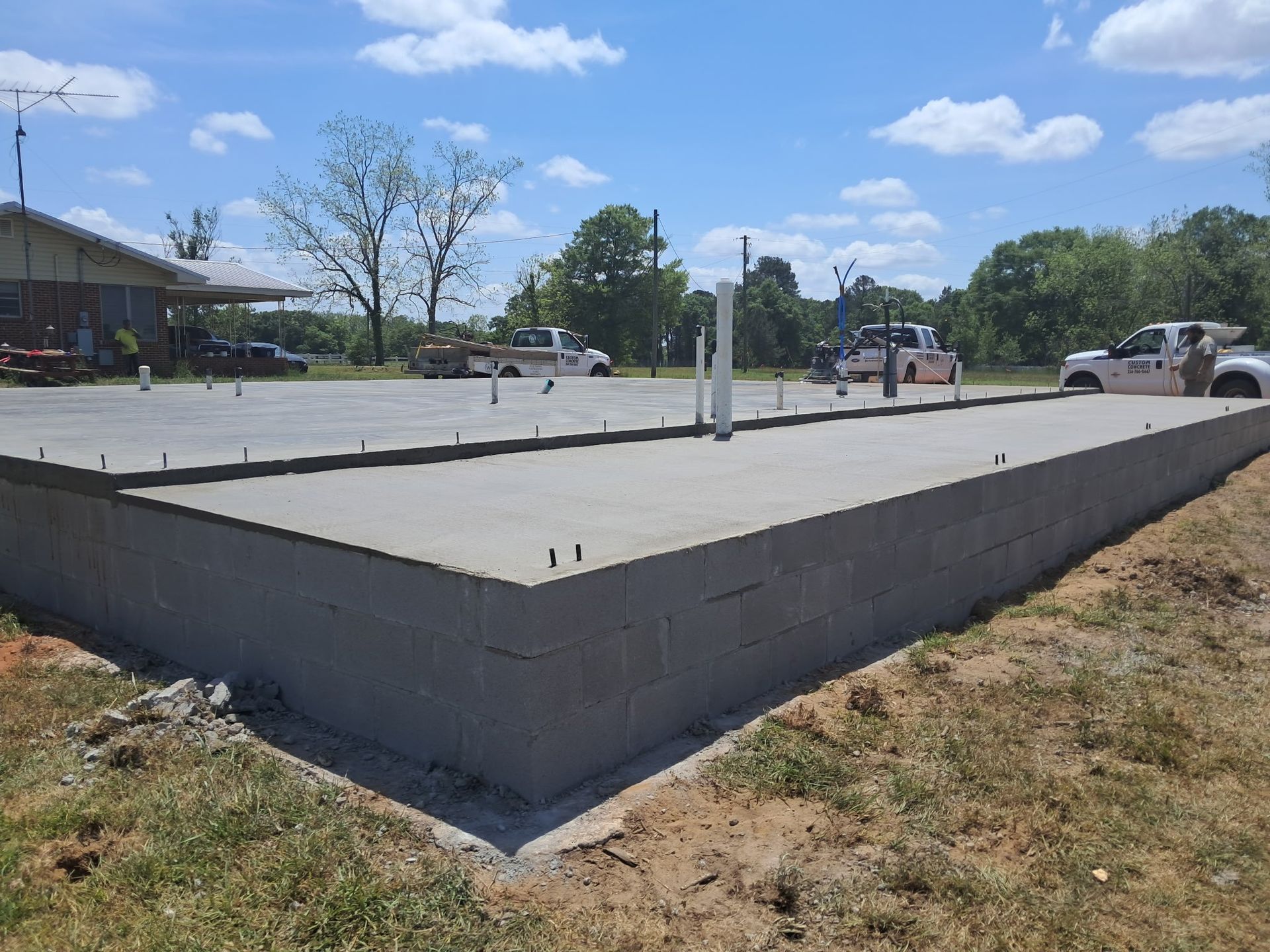 Newly poured concrete foundation with protruding rebar, in front of a building and trucks.