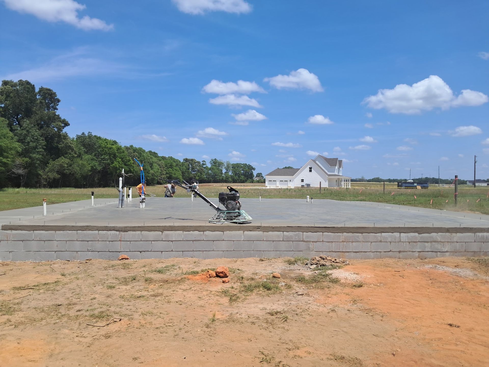 Construction site with a concrete slab in preparation, equipment, and a house in the distance under a blue sky.