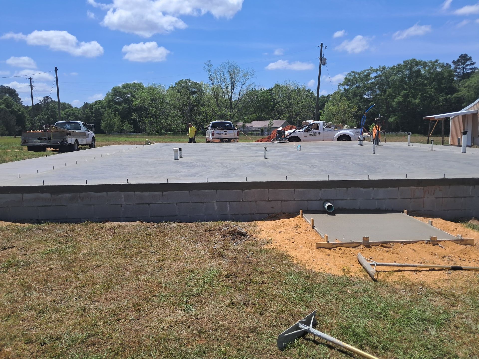 Concrete foundation slab under construction, outdoors on a sunny day. Workers and vehicles are in the background.