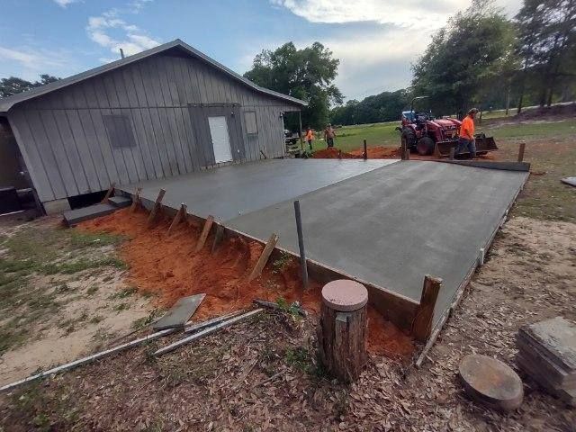 Newly poured concrete slab in front of a gray wooden building; a tractor is in the background.