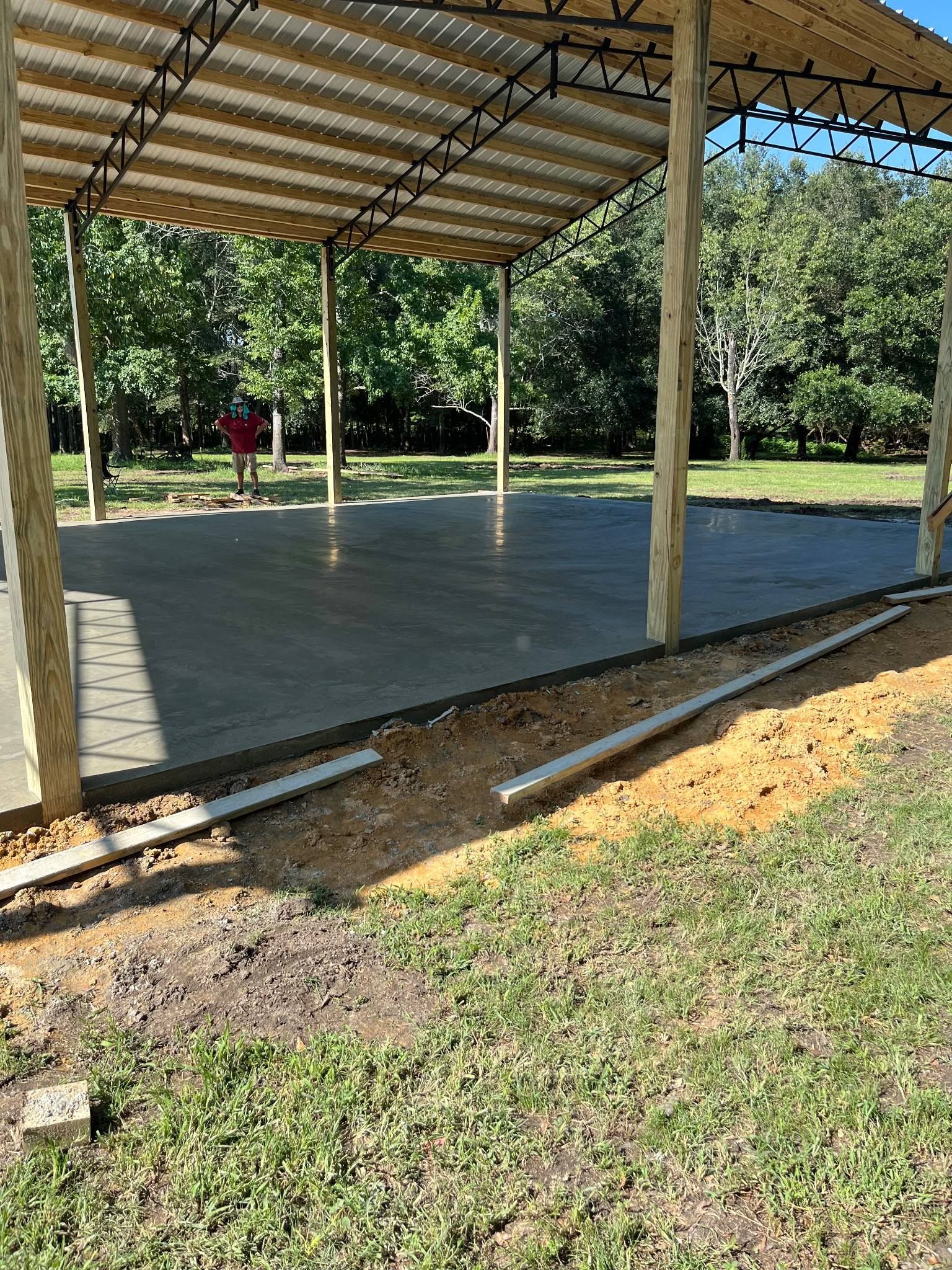 Newly poured concrete slab under a wooden-framed shelter with a corrugated roof. Person stands in the distance.
