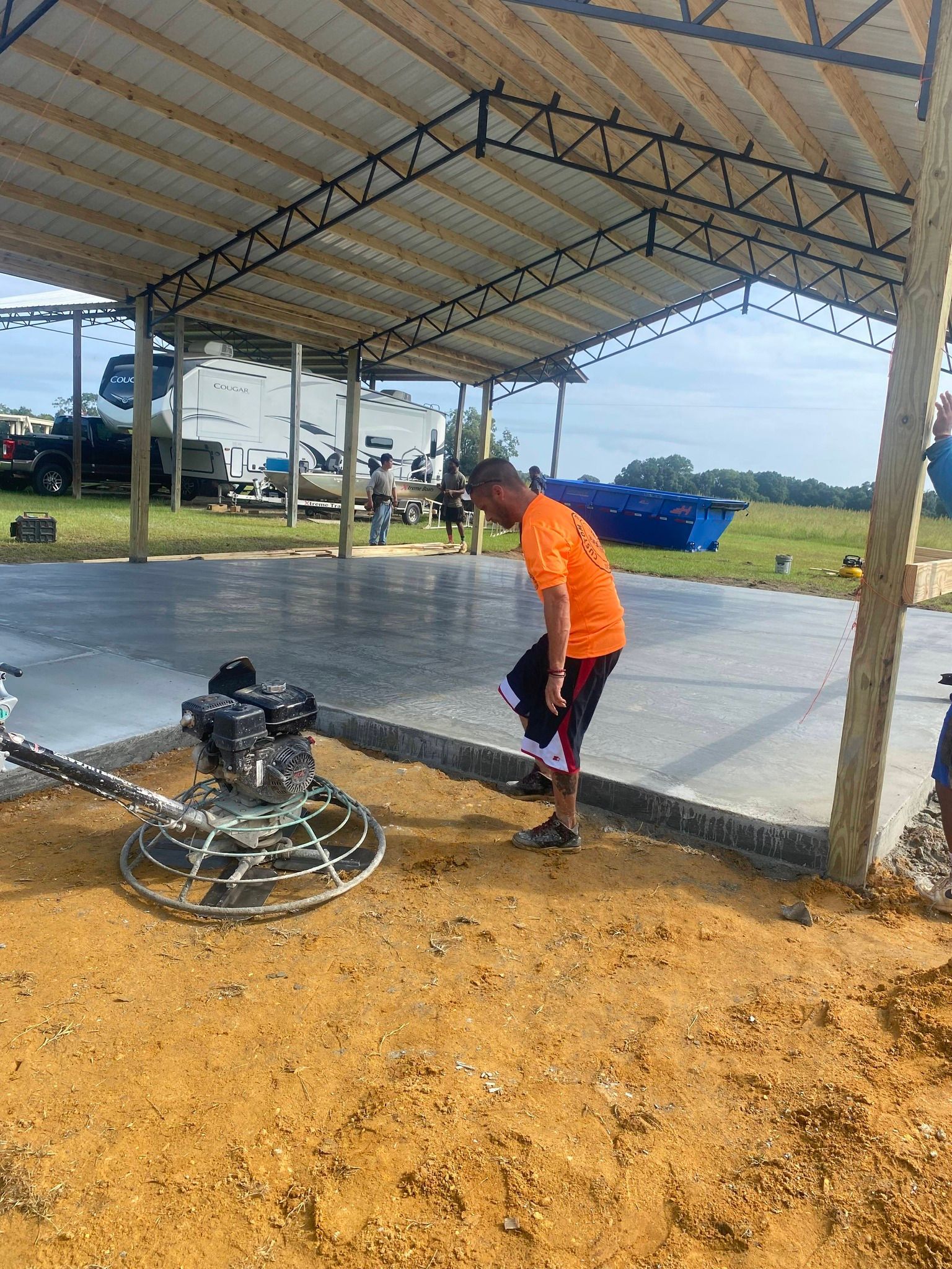 Man using a power trowel on a concrete slab under a metal roof structure.