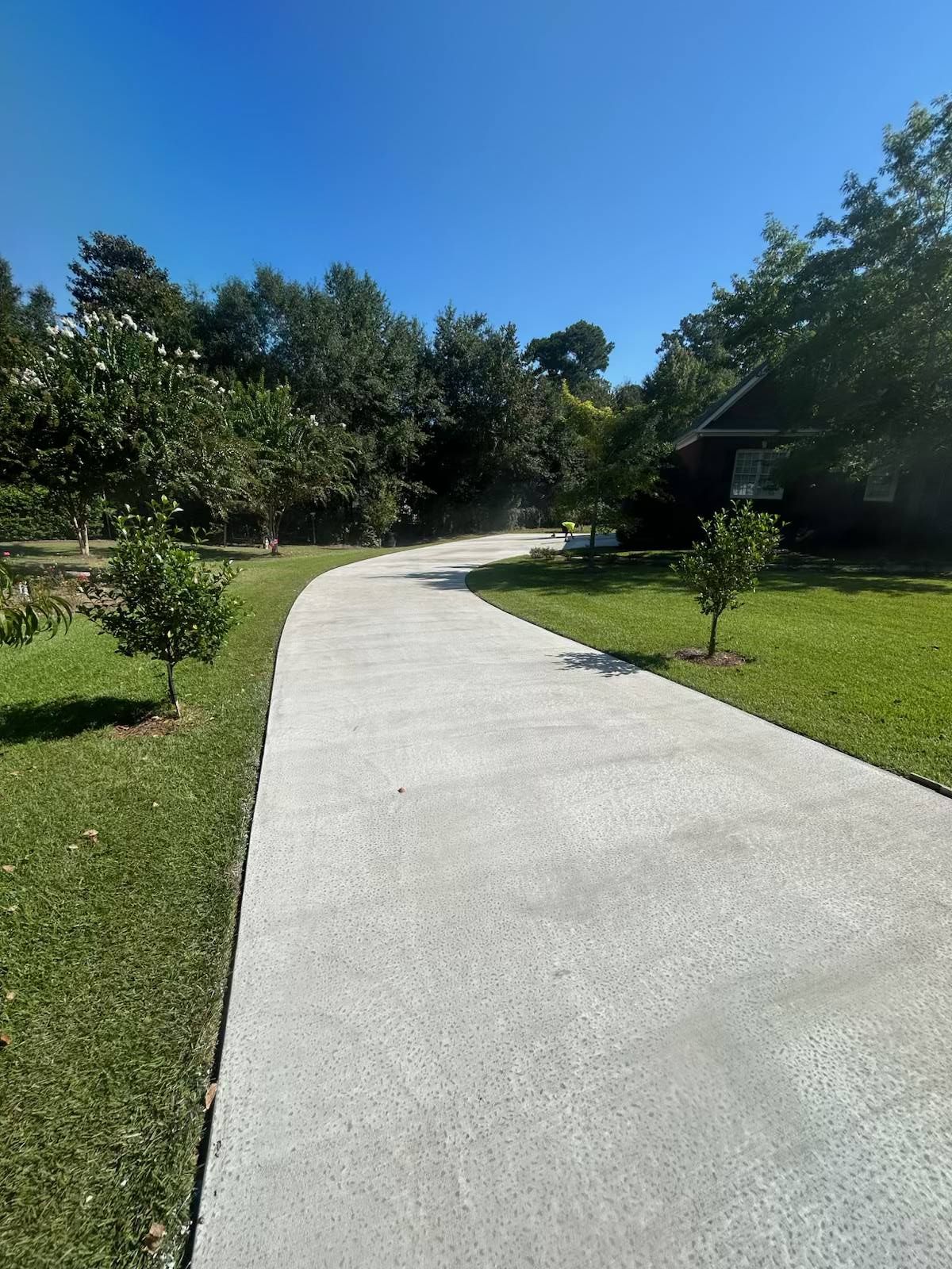 Concrete driveway curving towards a house, lined with grass and trees under a blue sky.