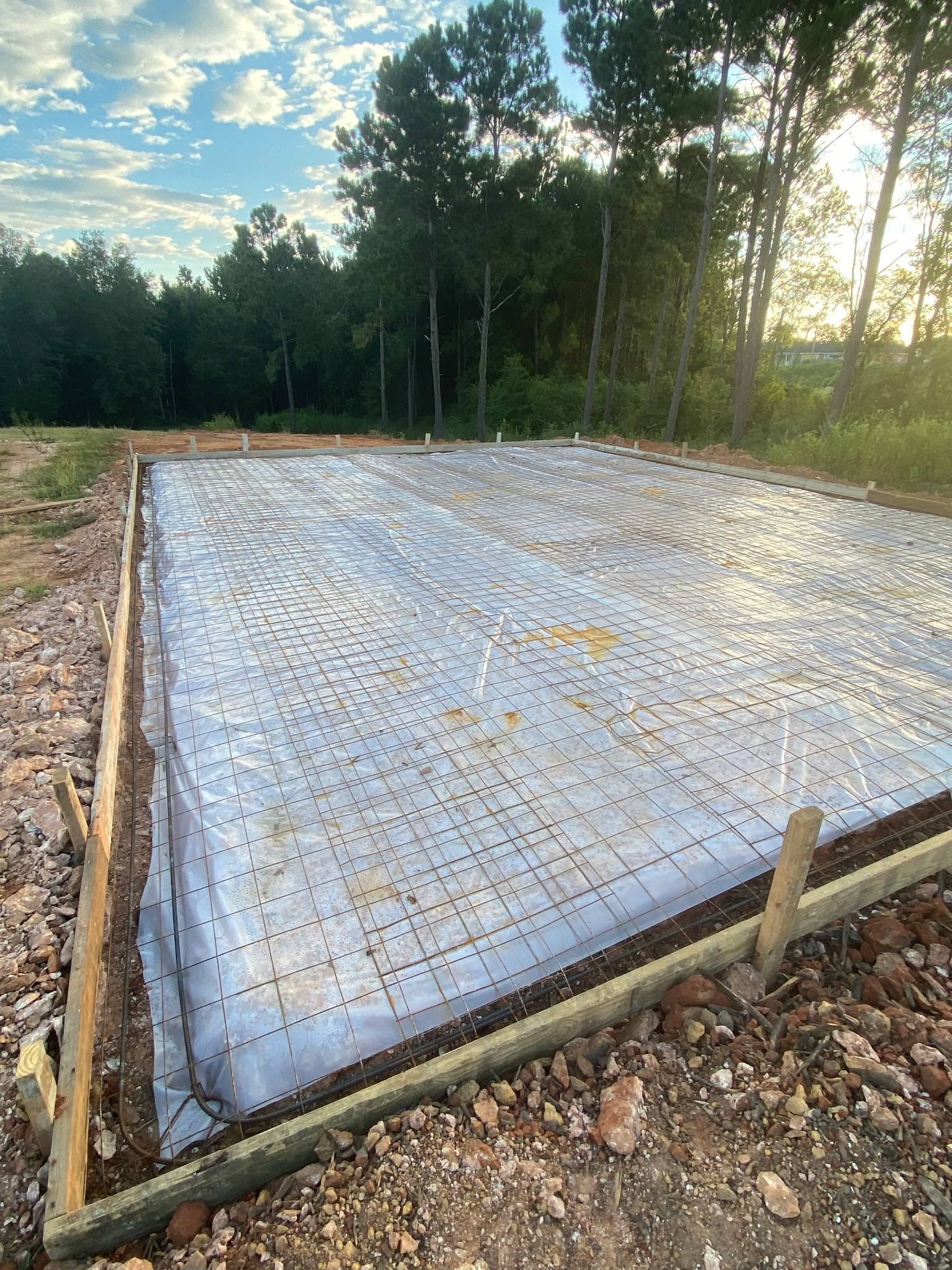 Foundation for a structure covered with plastic sheeting within wooden frame, surrounded by dirt and trees.