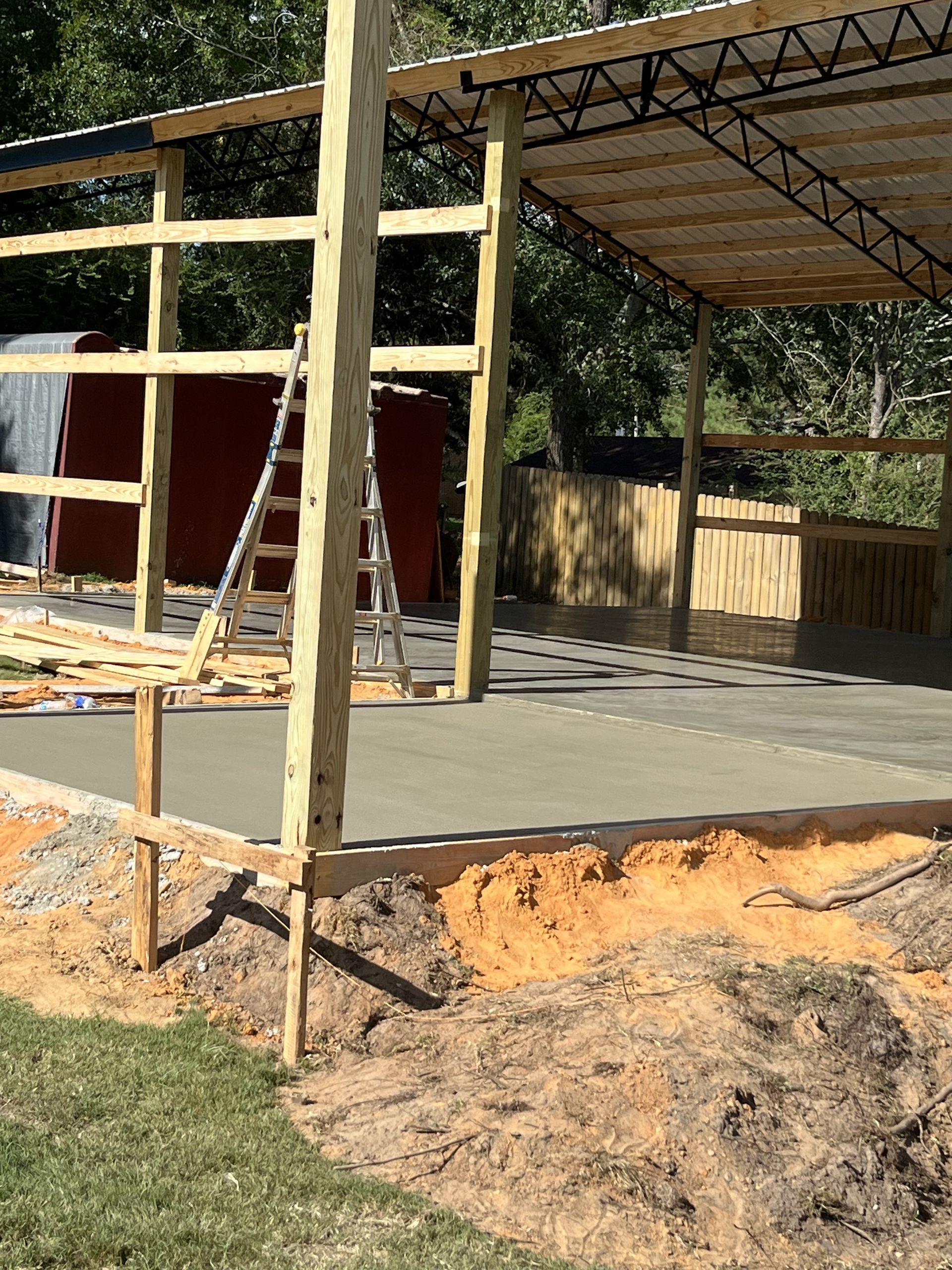 Construction site: concrete floor being poured under a metal roof frame. Wooden supports and ladder visible.