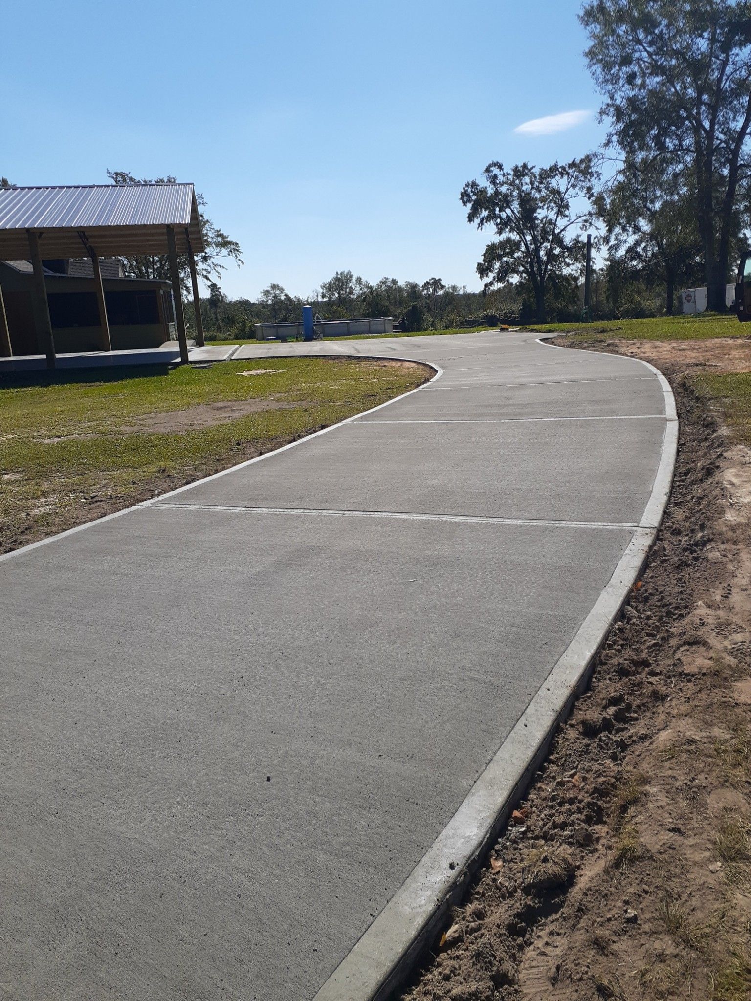 A paved pathway with white lines curves through a grassy area, near a building and trees under a blue sky.