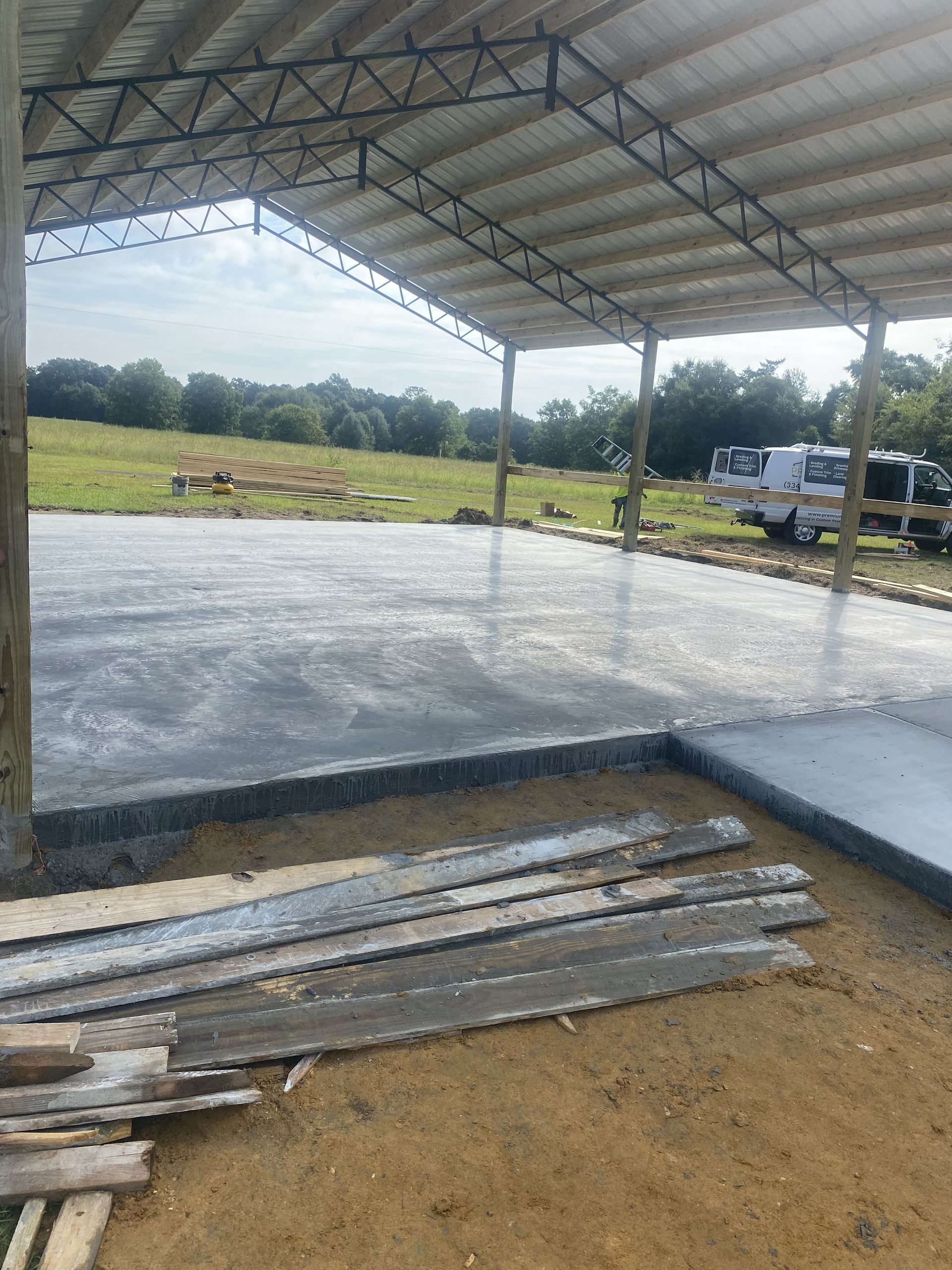 Newly poured concrete floor under a partially built metal roof, with a grassy field in the background.