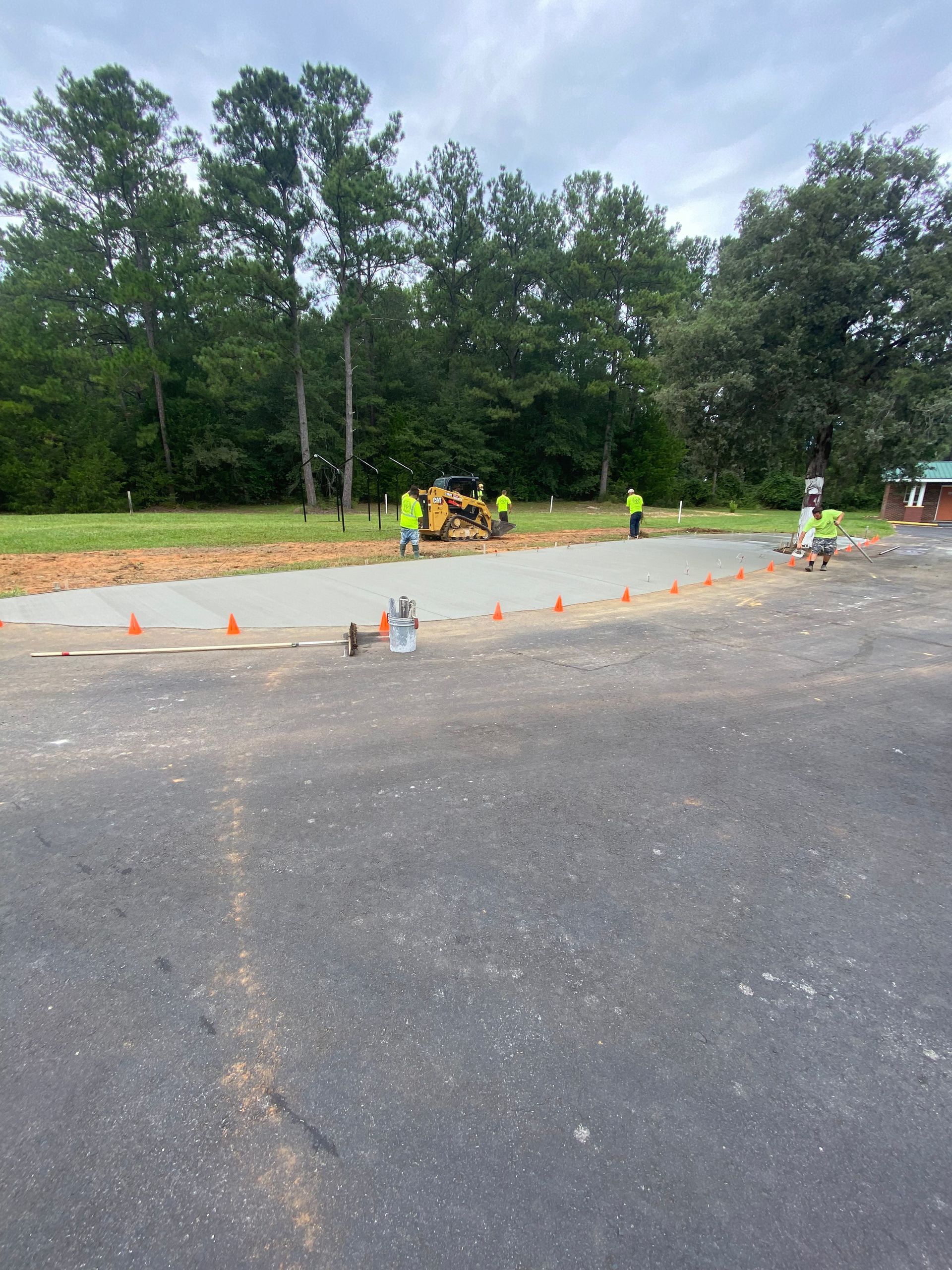 Construction workers paving a concrete section on a driveway near a tree line; orange cones delineate the area.