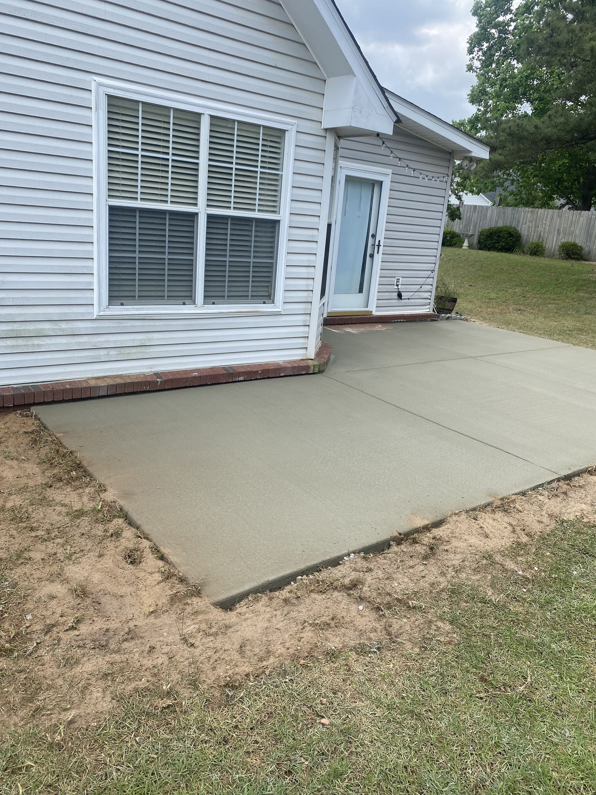Newly poured concrete patio next to a house with a window and door, surrounded by grass and dirt.