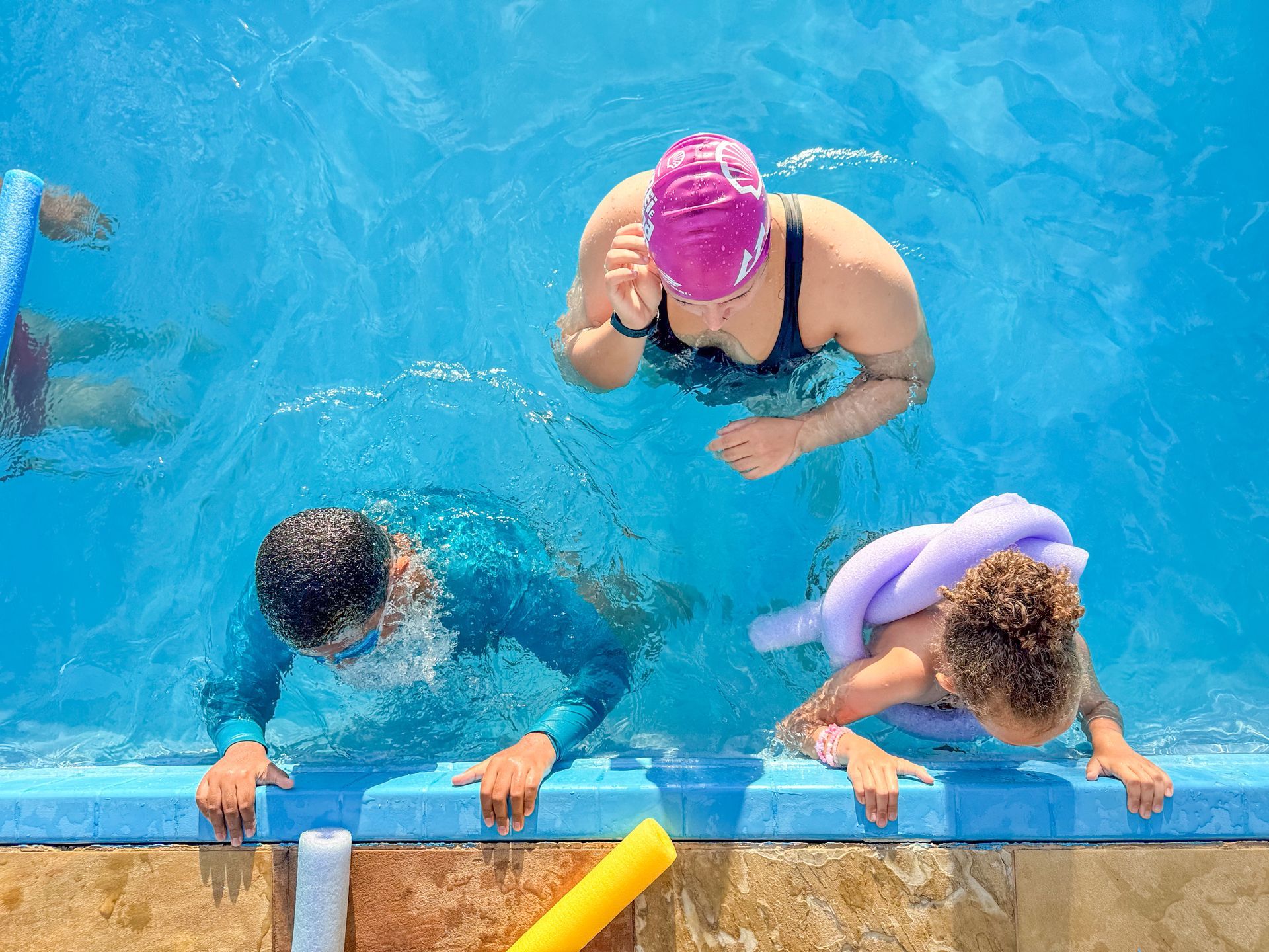 Pessoas em uma piscina. Uma mulher com uma touca rosa, uma pessoa com óculos de proteção e uma criança com uma boia.