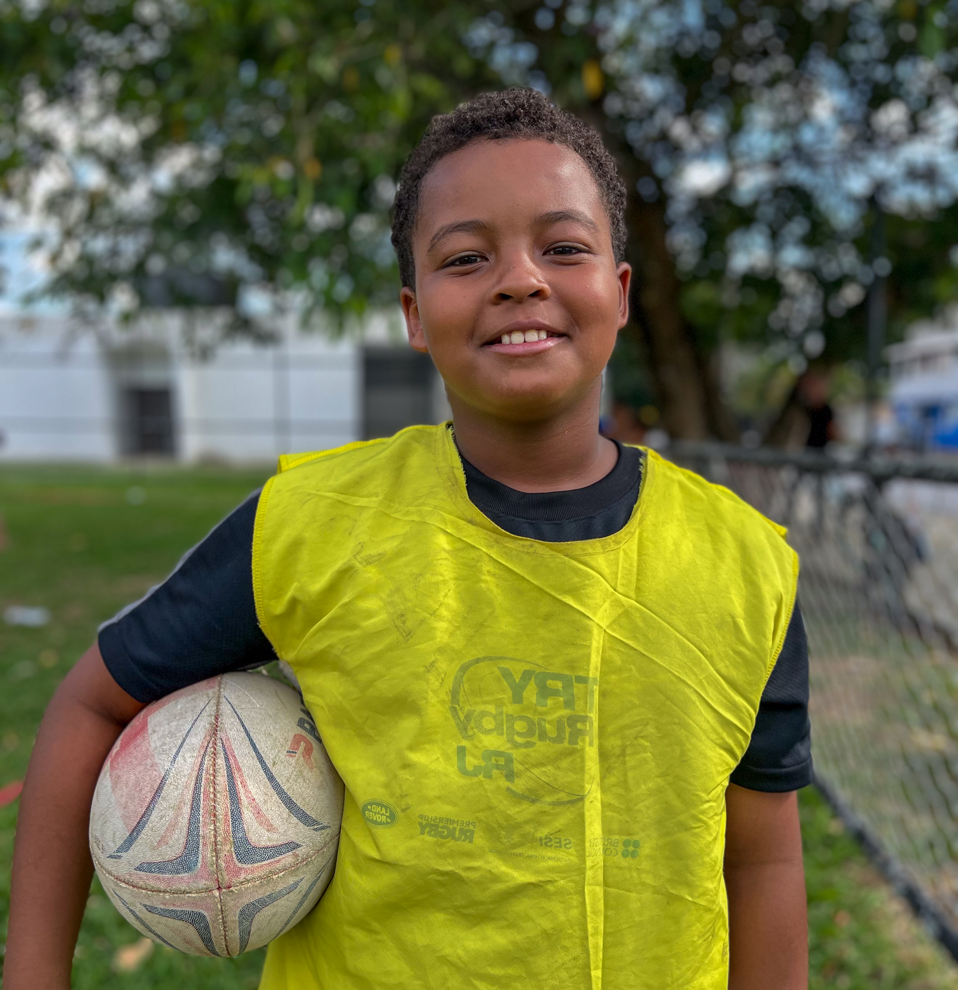 Menino de colete amarelo segurando uma bola de futebol, sorrindo ao ar livre.