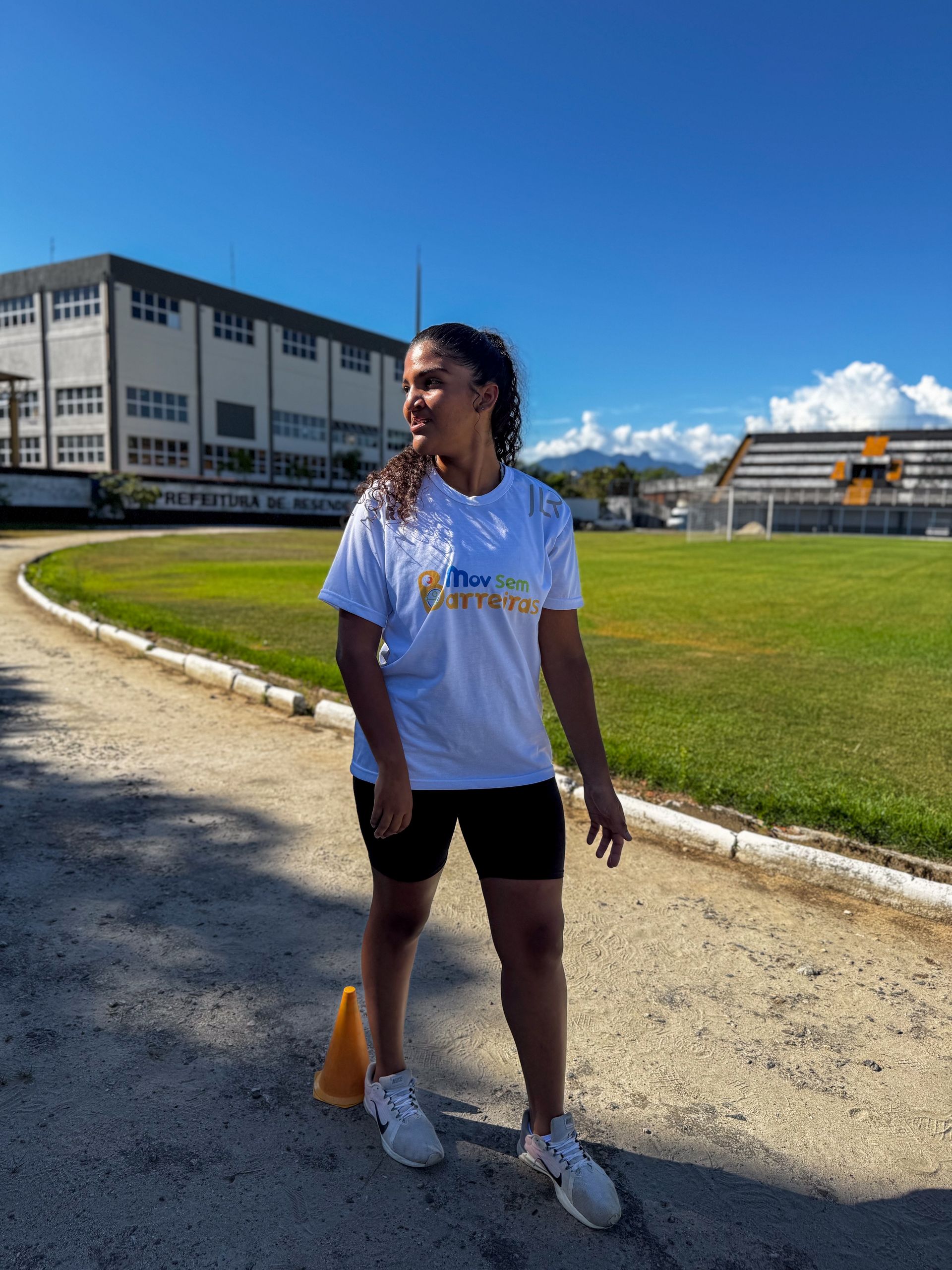 Mulher de camiseta branca e shorts preto em uma pista de corrida, olhando para o lado. Céu azul, prédio ao fundo.