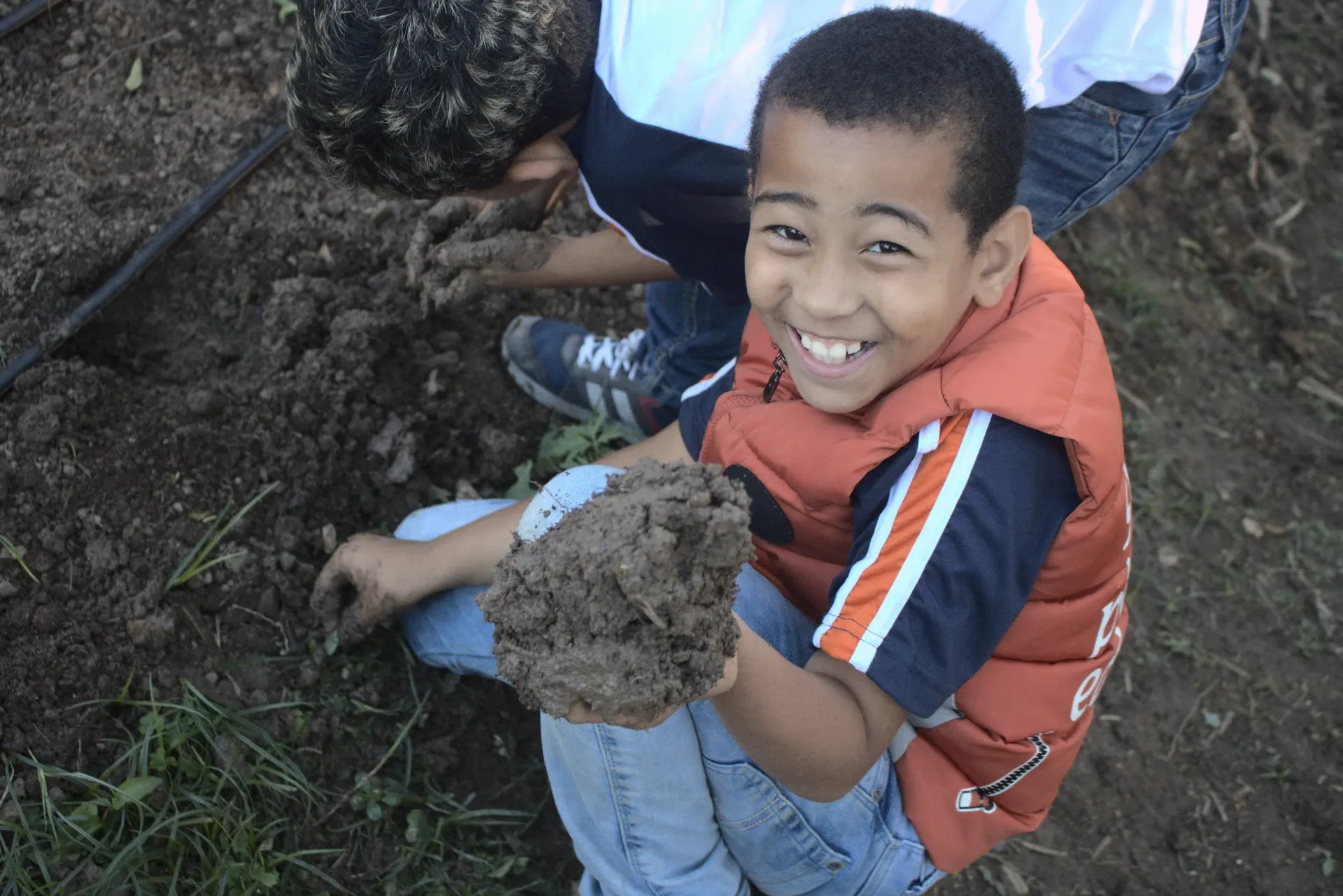 Menino de colete laranja sorri, segurando um punhado de terra enquanto faz jardinagem com um amigo.