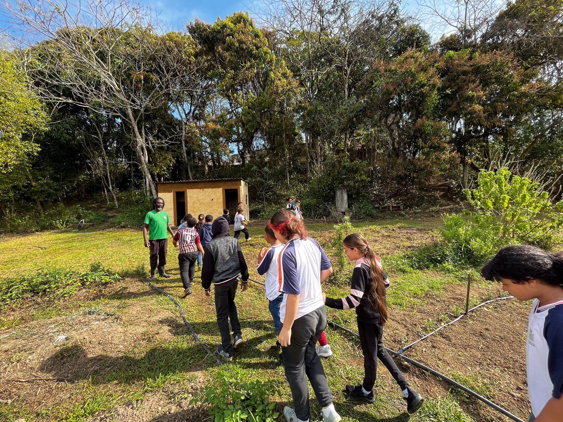 Crianças caminhando em um campo gramado em direção a um pequeno prédio em frente às árvores.