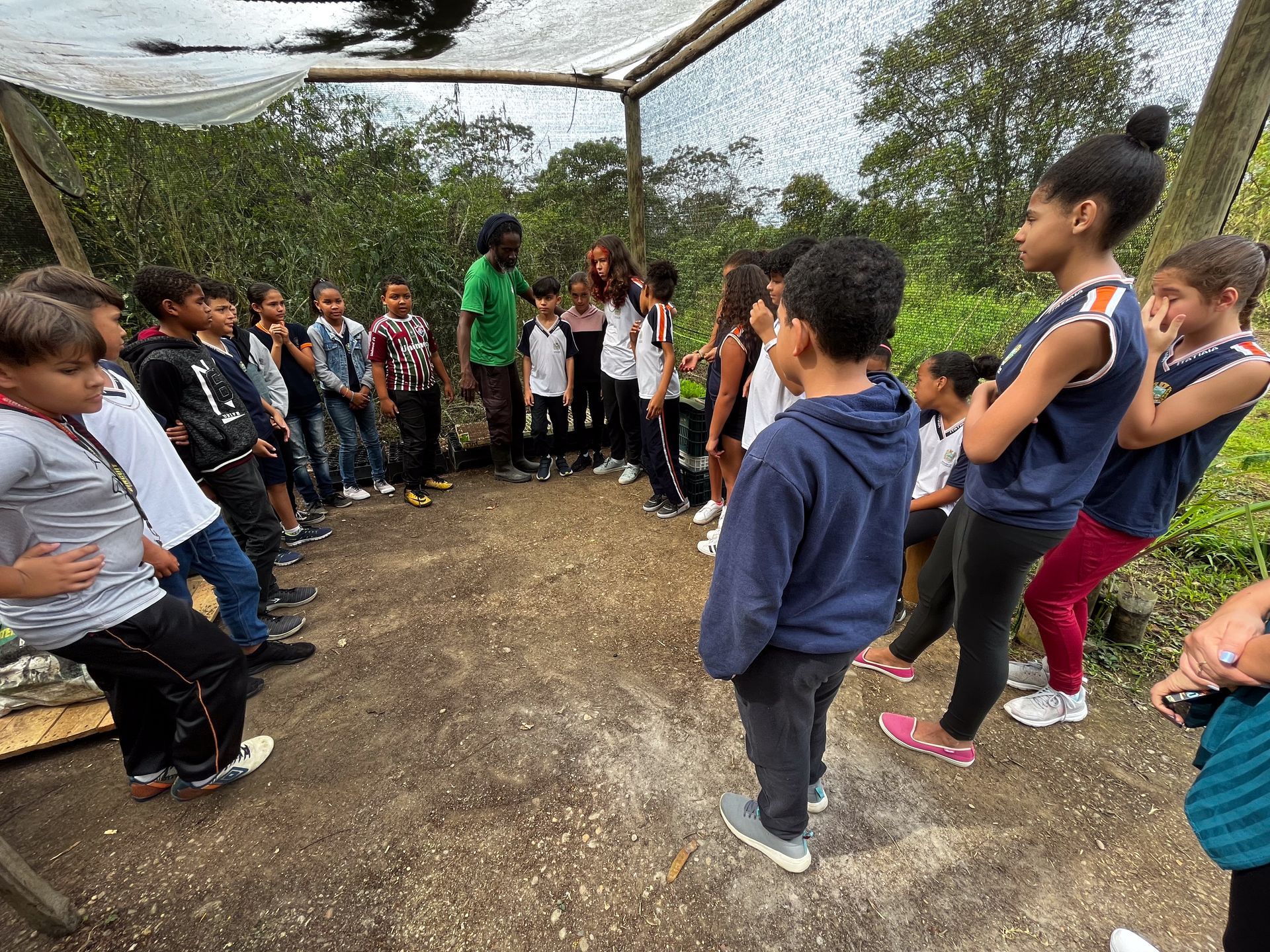 Crianças e professora em círculo ao ar livre, sob uma rede. Algumas olham para algo no centro.